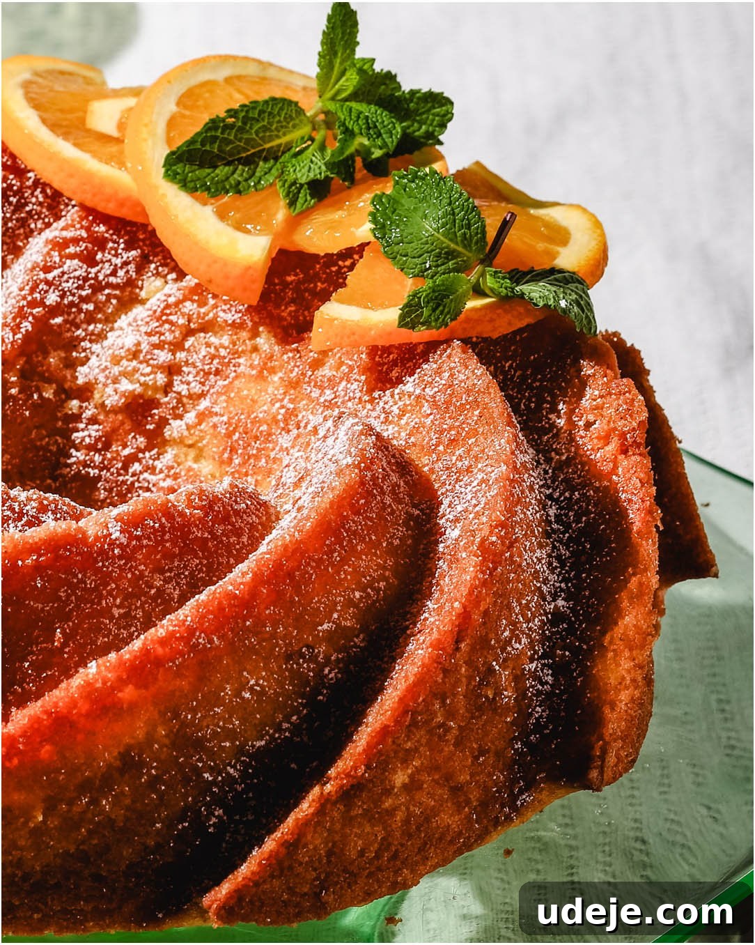 Delicate Orange Cardamom Cake 3 Beautifully baked orange colored bundt cake, lightly dusted with powdered sugar, and elegantly garnished with fresh mint leaves and vibrant orange slices, presented on a striking green glass cake stand.