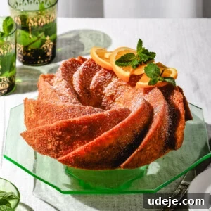 Orange colored bundt cake topped with powdered sugar, orange slices and mint leaves on a green octagon shape glass cake stand, with green and gold glasses with water and mint leaves, on a white table cloth with a cake knife and white plates at the side.