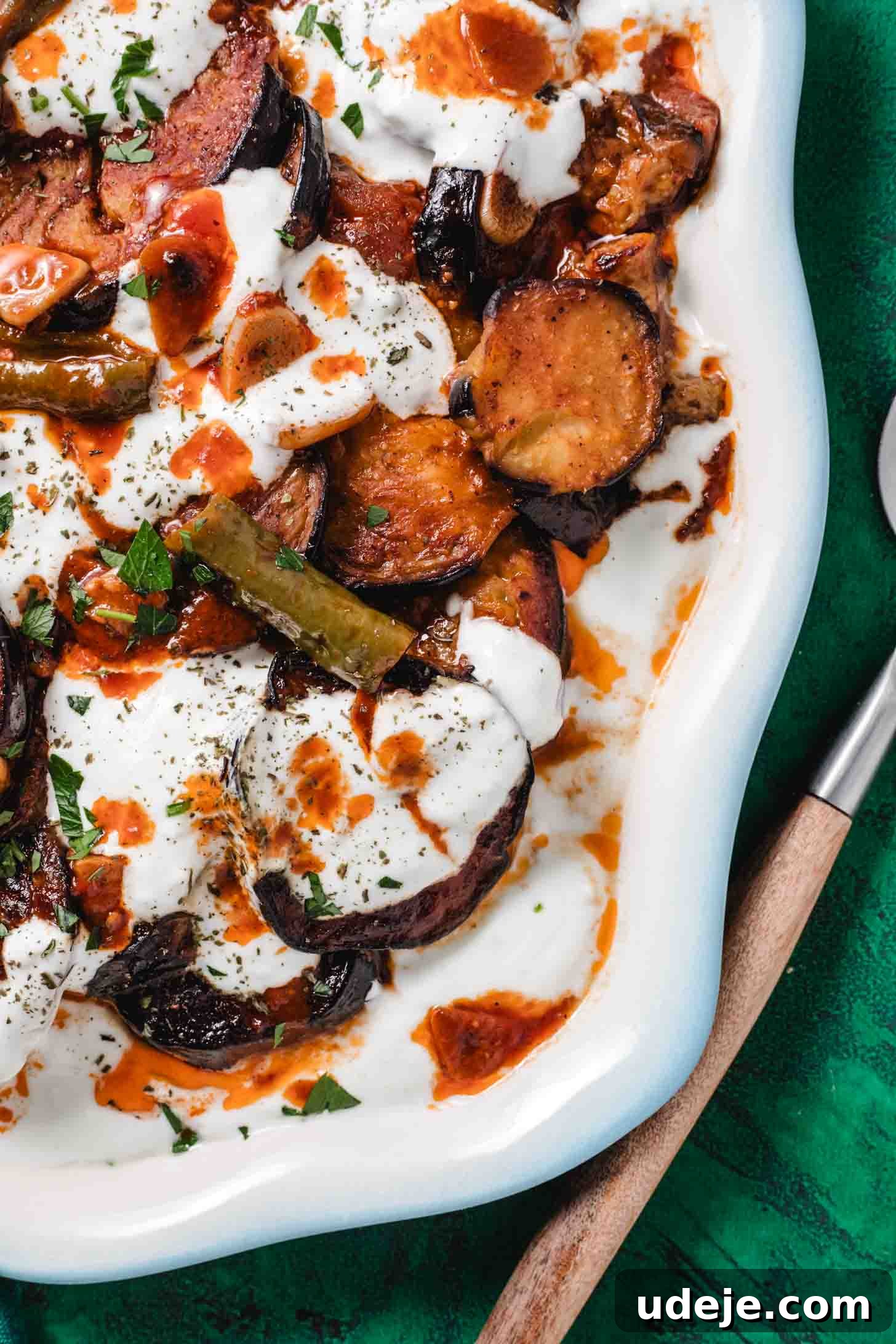Borani Banjan Creamy Afghan Eggplant Yogurt Dip 13 A close-up view of Borani Banjan in a pristine white dish, showcasing an enticing arrangement of fried eggplant slices meticulously layered with creamy yogurt, a rich spiced tomato sauce, fresh green peppers, and a delicate sprinkling of herbs. The dish is artfully placed on a vibrant green surface, with a wooden-handled spoon resting beside it, inviting a taste of this popular Afghan Eggplant and yogurt dip.