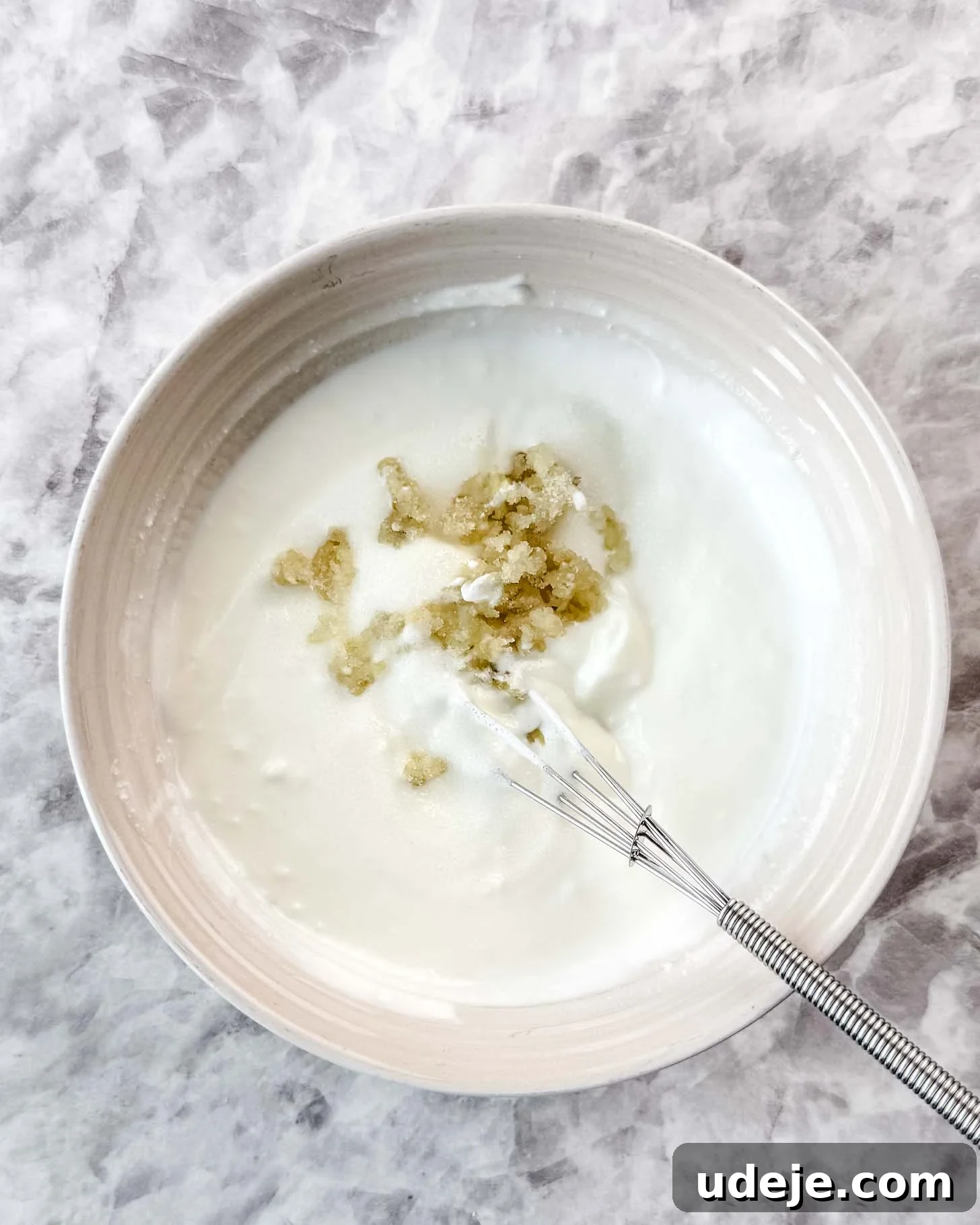 Borani Banjan Creamy Afghan Eggplant Yogurt Dip 4 A creamy bowl of yogurt, subtly enhanced with grated ginger and a touch of sugar, sits elegantly on a light marble surface. A small whisk rests in the bowl, suggesting gentle preparation for this Borani Banjan inspired sauce.