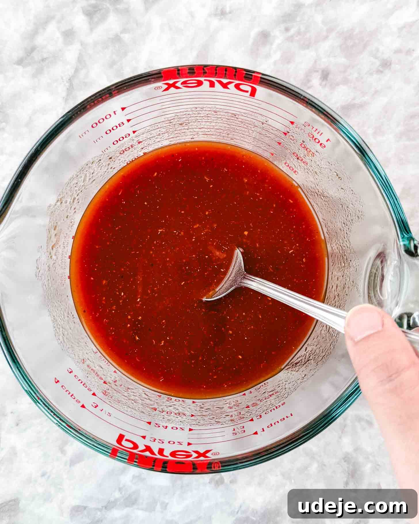 Borani Banjan Creamy Afghan Eggplant Yogurt Dip 5 A hand gently stirs a rich, reddish-brown Borani Banjan sauce in a clear glass Pyrex measuring cup, resting on a clean, light marble surface. The vibrant color of the sauce hints at its delicious, spiced tomato base.