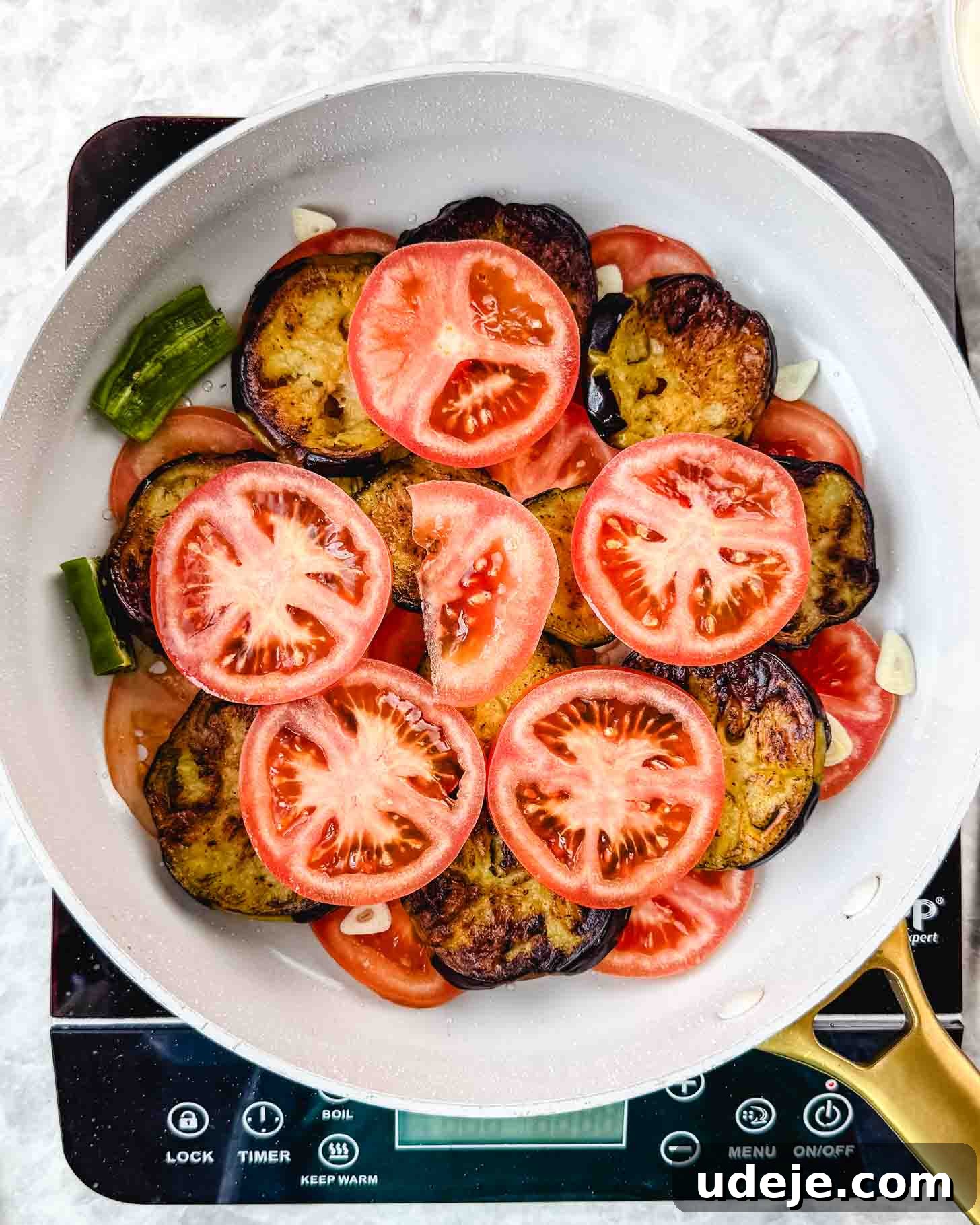 Borani Banjan Creamy Afghan Eggplant Yogurt Dip 9 A white skillet on an induction cooktop is filled with vibrant layers of sliced tomatoes, golden-brown eggplant rounds, bright green pepper pieces, and fragrant garlic cloves. This artful arrangement is simmering gently, ready for cooking this flavorful Borani Banjan.