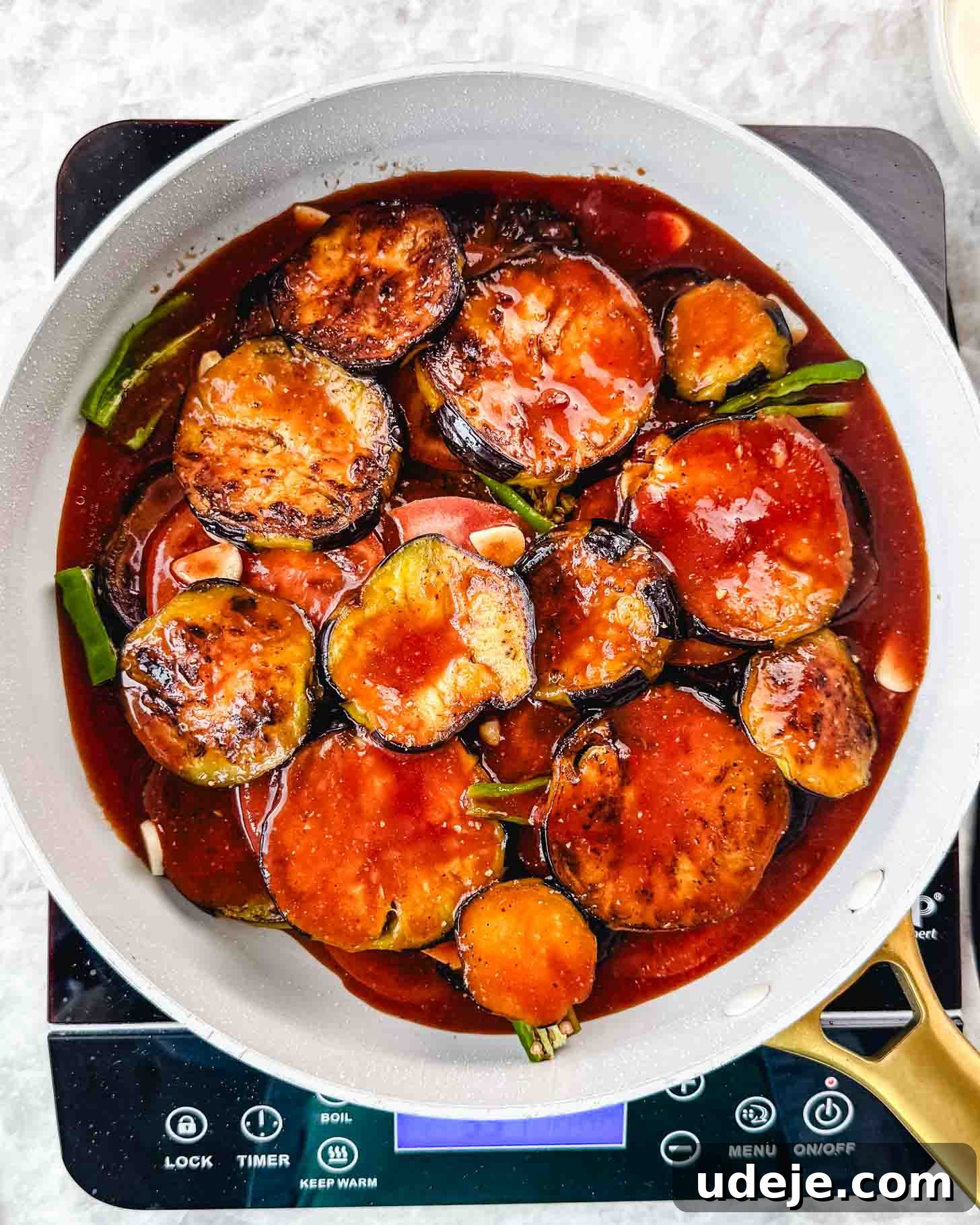 Borani Banjan Creamy Afghan Eggplant Yogurt Dip 10 A white skillet sits on a stovetop, filled with tender eggplant slices coated in a glossy, rich red sauce, garnished with fresh green onions and delicate garlic slices. The simmering dish, reminiscent of Borani Banjan, exudes warmth and promises an authentic Afghan culinary experience.