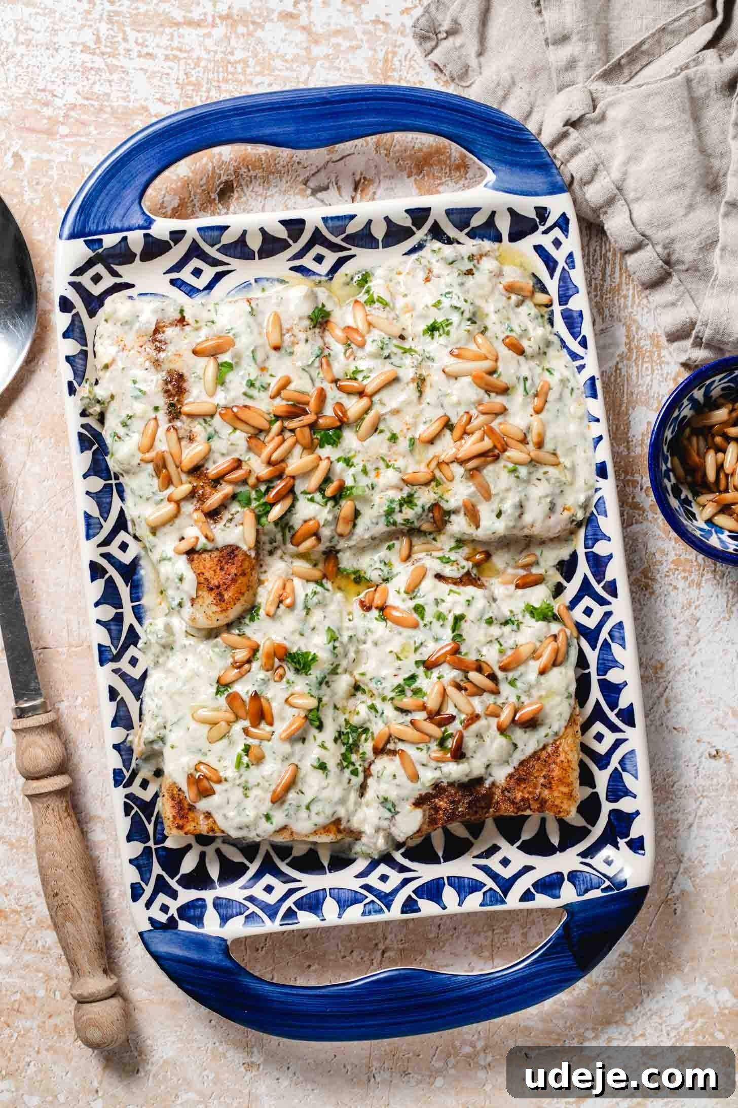 Samke Harra: Lebanon's Signature Spicy Fish 2 Pieces of golden fish smothered in a white sauce with golden pine nuts and chopped parsley on a blue patterned tray with handles, a beige dish towel, a small blue patterned bowl with pine nuts and a wooden handled serving spoon at the side on a beige and white background.