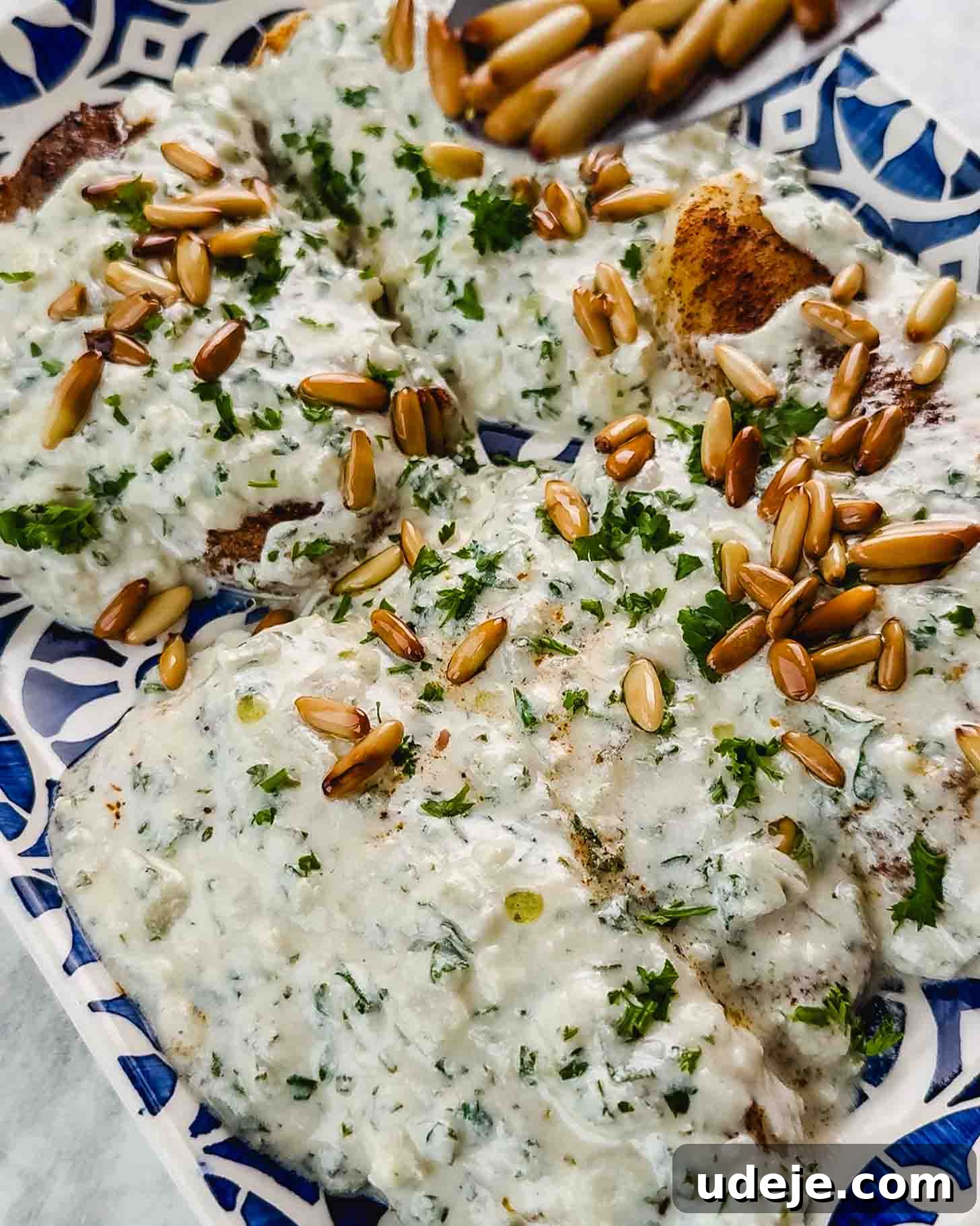 Samke Harra: Lebanon's Signature Spicy Fish 9 Pieces of golden fish smothered in a white sauce with golden pine nuts being sprinkled on top and chopped parsley on a blue patterned tray.