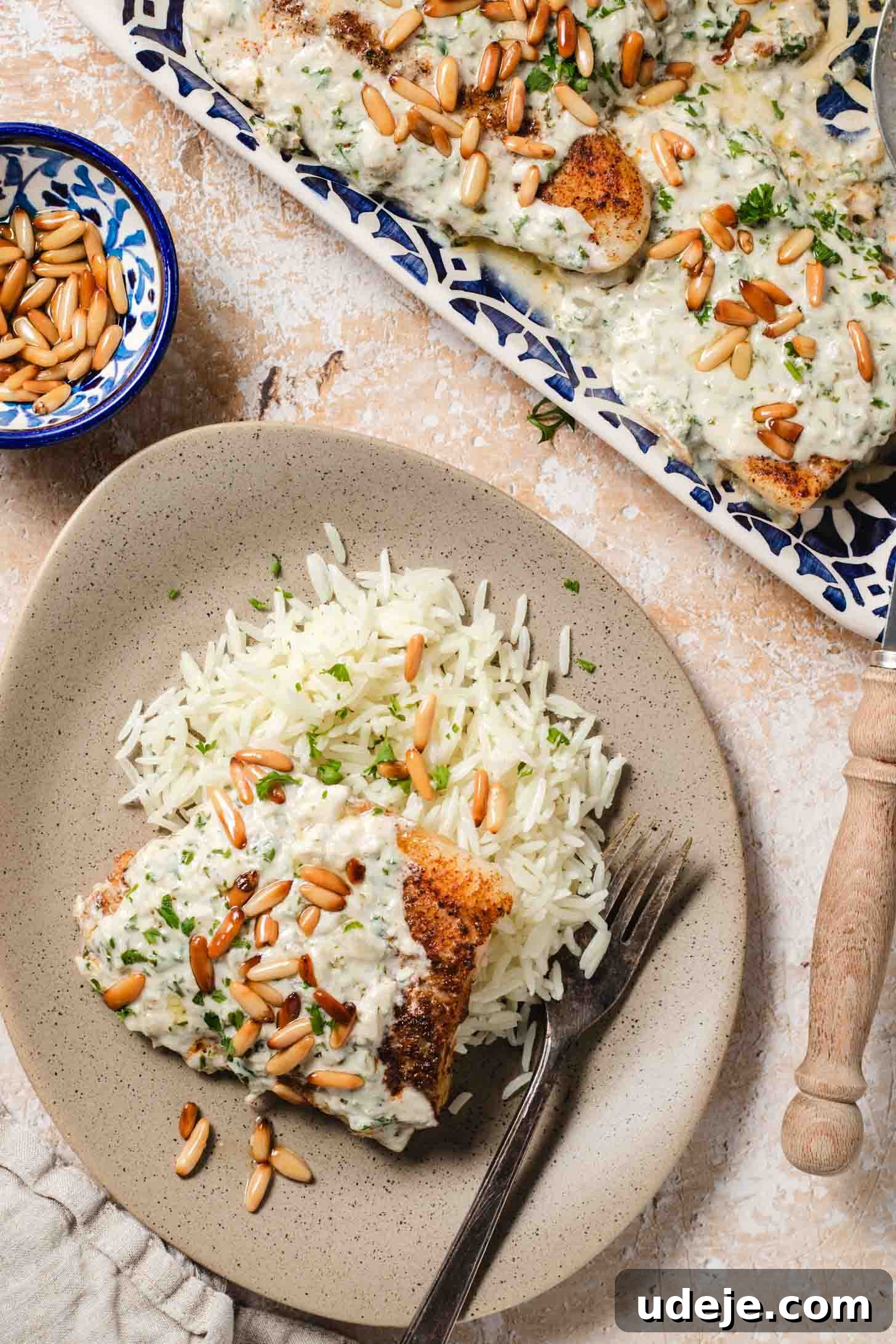 Samke Harra: Lebanon's Signature Spicy Fish 10 Pieces of golden fish smothered in a white sauce with golden pine nuts and chopped parsley on a blue patterned tray with handles, a beige plate with rice a piece of golden fish with white sauce and golden pine nuts, a beige dish towel, a small blue patterned bowl with pine nuts and a wooden handled serving spoon at the side on a beige and white background.