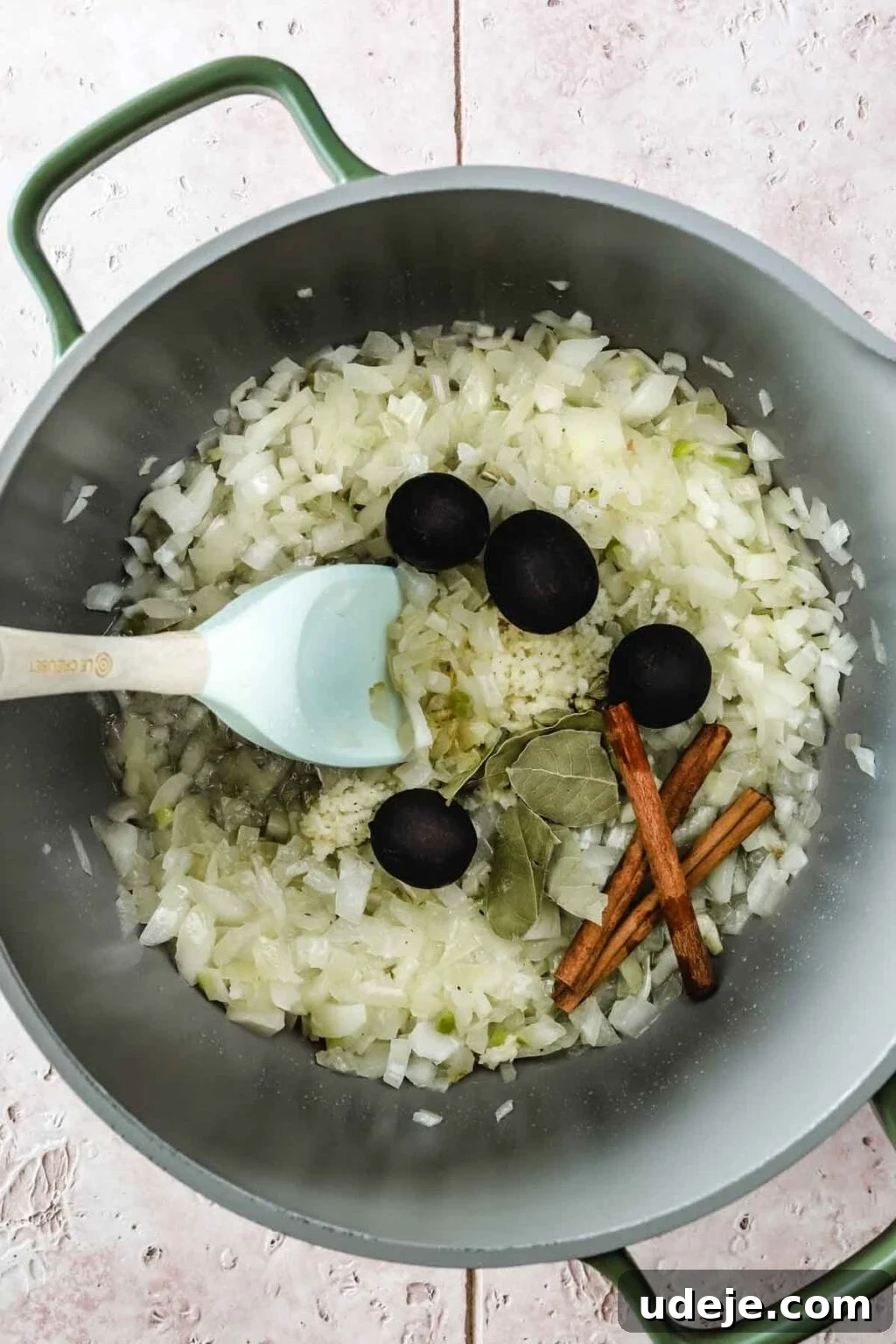 Machboos and Kabsa Aromatic Chicken and Rice 5 Large grey pot with green handles with chopped onions, dry limes, bay leaves and cinnamon sticks with a light blue spoon on a light coloured tiled background
