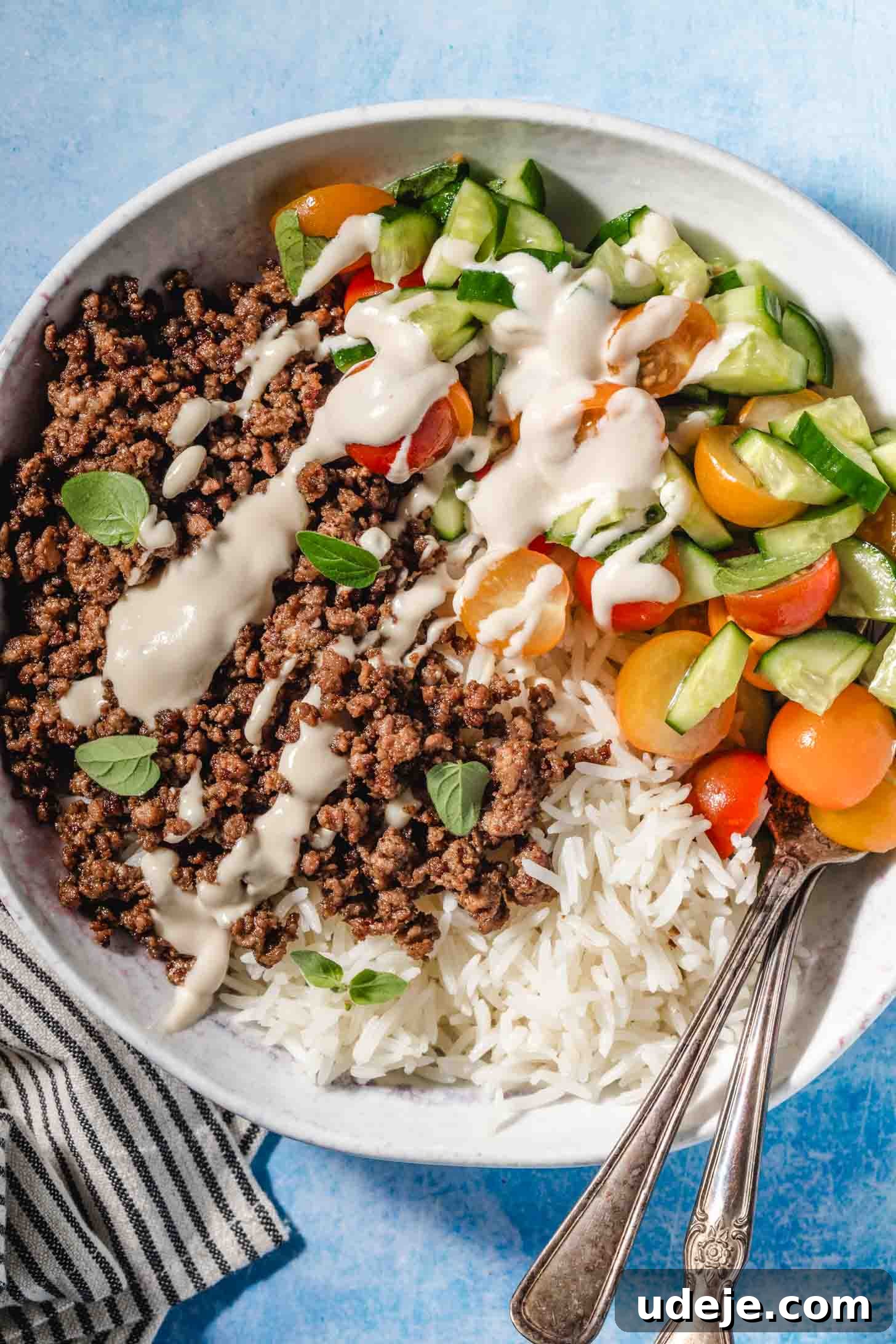 Easy Middle Eastern Spiced Beef Bowls 2 A vibrant Middle Eastern Ground Beef Bowl with perfectly cooked, seasoned ground beef, fluffy white rice, a fresh chopped salad featuring lettuce, halved cherry tomatoes, and cucumbers. A creamy beige tahini sauce is drizzled over the top, ready to be enjoyed. Two spoons rest beside the bowl on a striped tea towel against a blue background.
