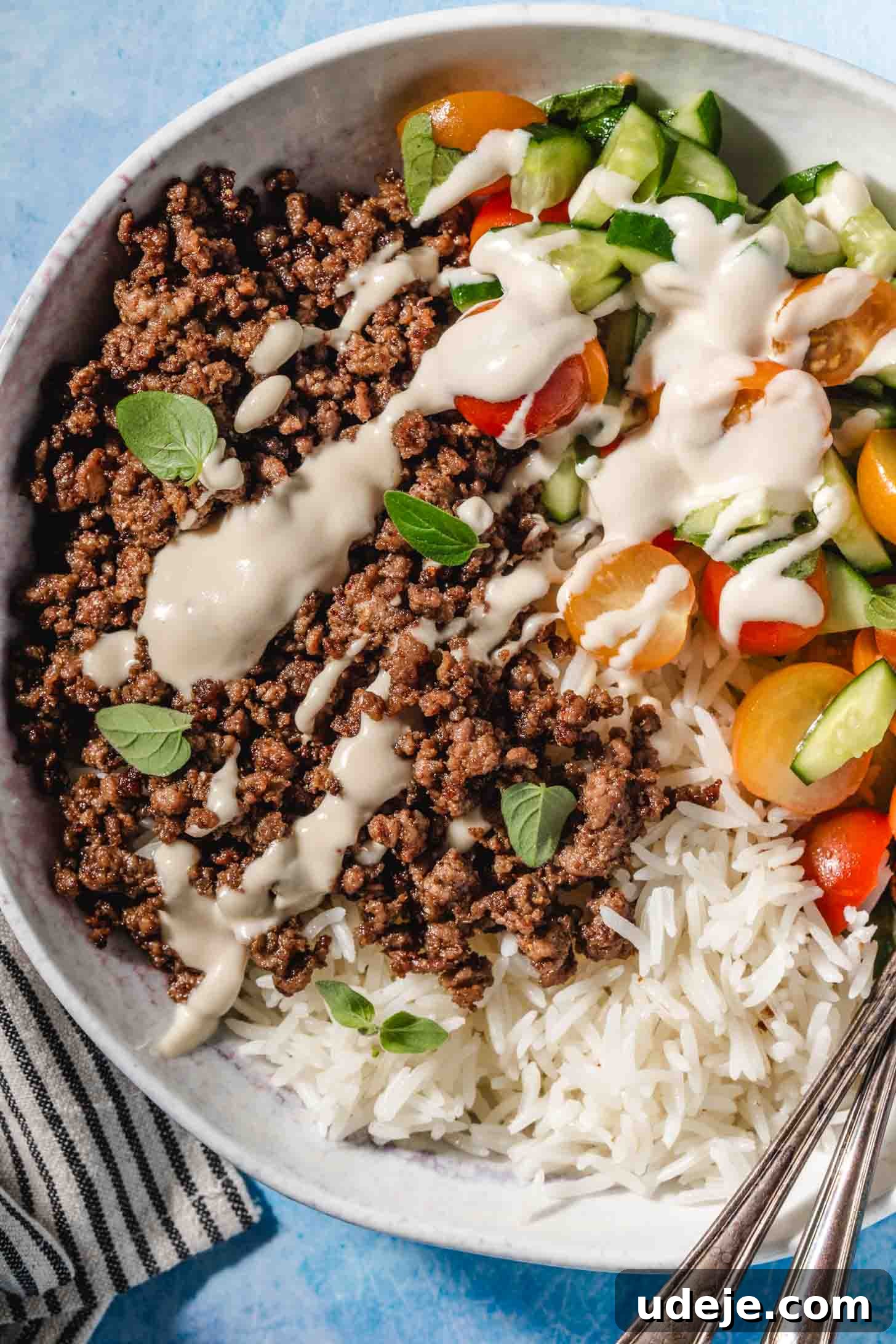 Easy Middle Eastern Spiced Beef Bowls 6 A beautifully composed Middle Eastern Ground Beef Dinner Bowl, featuring well-seasoned, crispy ground beef, fluffy white rice, a crisp chopped salad, and a generous drizzle of creamy tahini sauce, presented on a blue background.