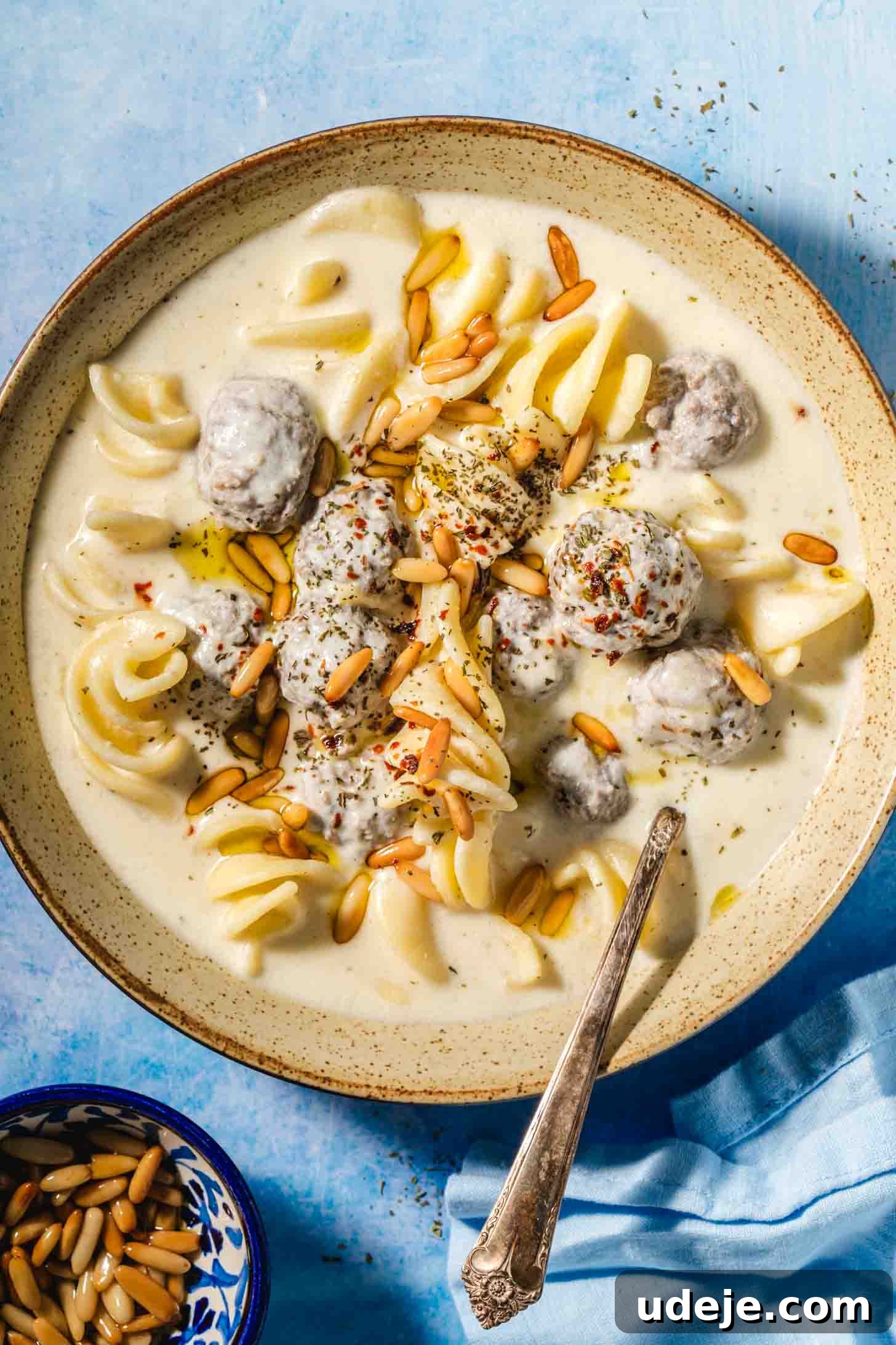 The Ultimate Meatball Soup with Yogurt and Pasta 2 A beige speckled bowl brimming with creamy, off-white liquid. Small, perfectly browned meatballs are submerged in the soup alongside spiral-shaped white pasta. Golden toasted pine nuts are scattered generously on top. A metal spoon rests in the bowl, all presented on a vibrant blue background with a matching blue tea towel in the corner and a small blue bowl of pine nuts.