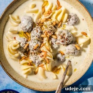 The Ultimate Meatball Soup with Yogurt and Pasta 9 Beige speckled bowl with a creamy off white liquid, small brown meatballs covered in the off white liquid, spiral shaped white pasta and golden toasted pine nuts on top with a metal spoon in the bowl on a blue background with a blue tea towel in the corner and a small blue bowl with toasted pine nuts.