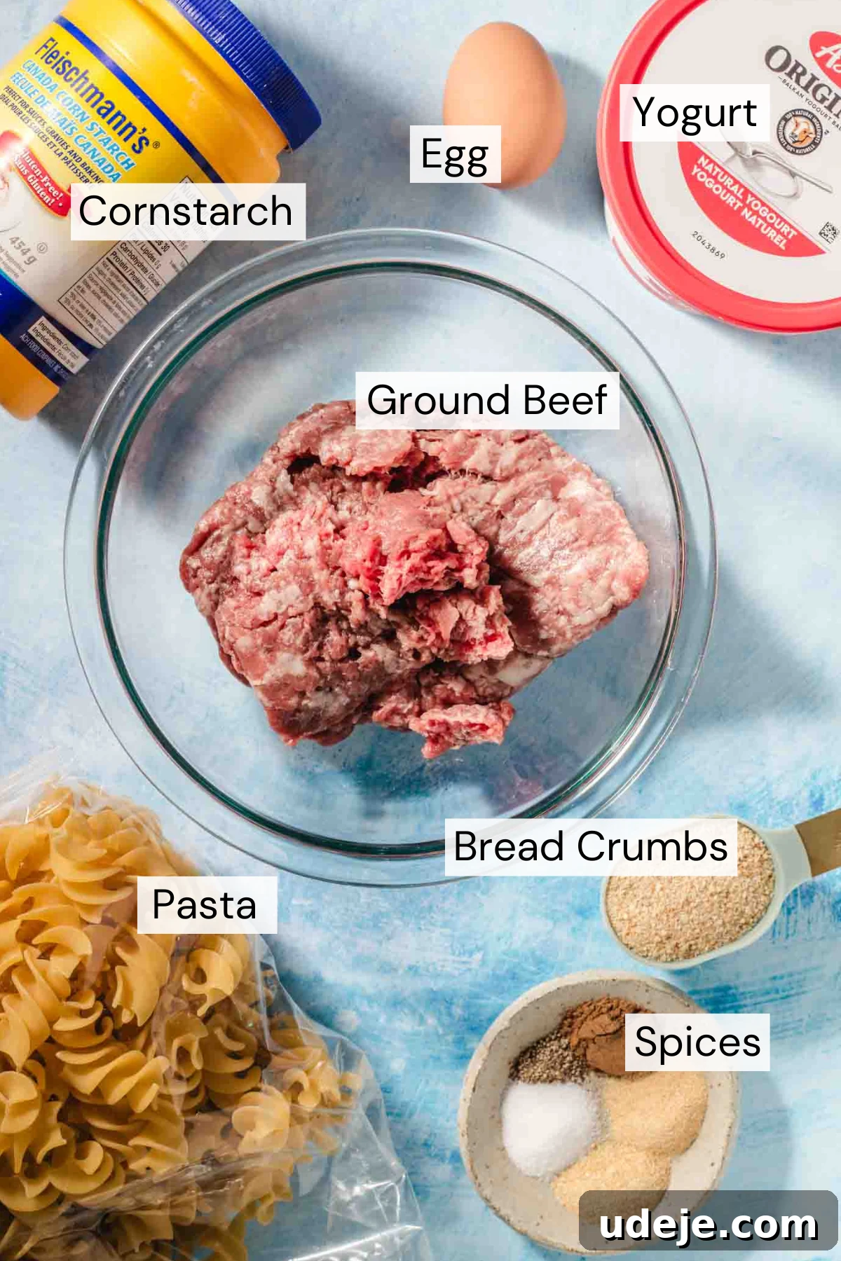 The Ultimate Meatball Soup with Yogurt and Pasta 3 A vibrant flat lay of ingredients for the soup. A large container of whole milk yogurt with a red lid sits beside a brown egg and a yellow and blue box of cornstarch. A glass bowl holds lean ground beef. A clear plastic package contains spiral pasta. A measuring spoon displays breadcrumbs, and a small bowl holds a blend of spices, all arranged on a bright blue background.