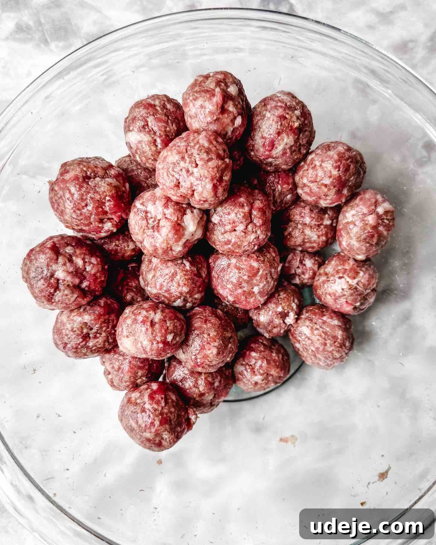 The Ultimate Meatball Soup with Yogurt and Pasta 4 Round brown uncooked meatballs piled into a glass bowl on a marble surface.