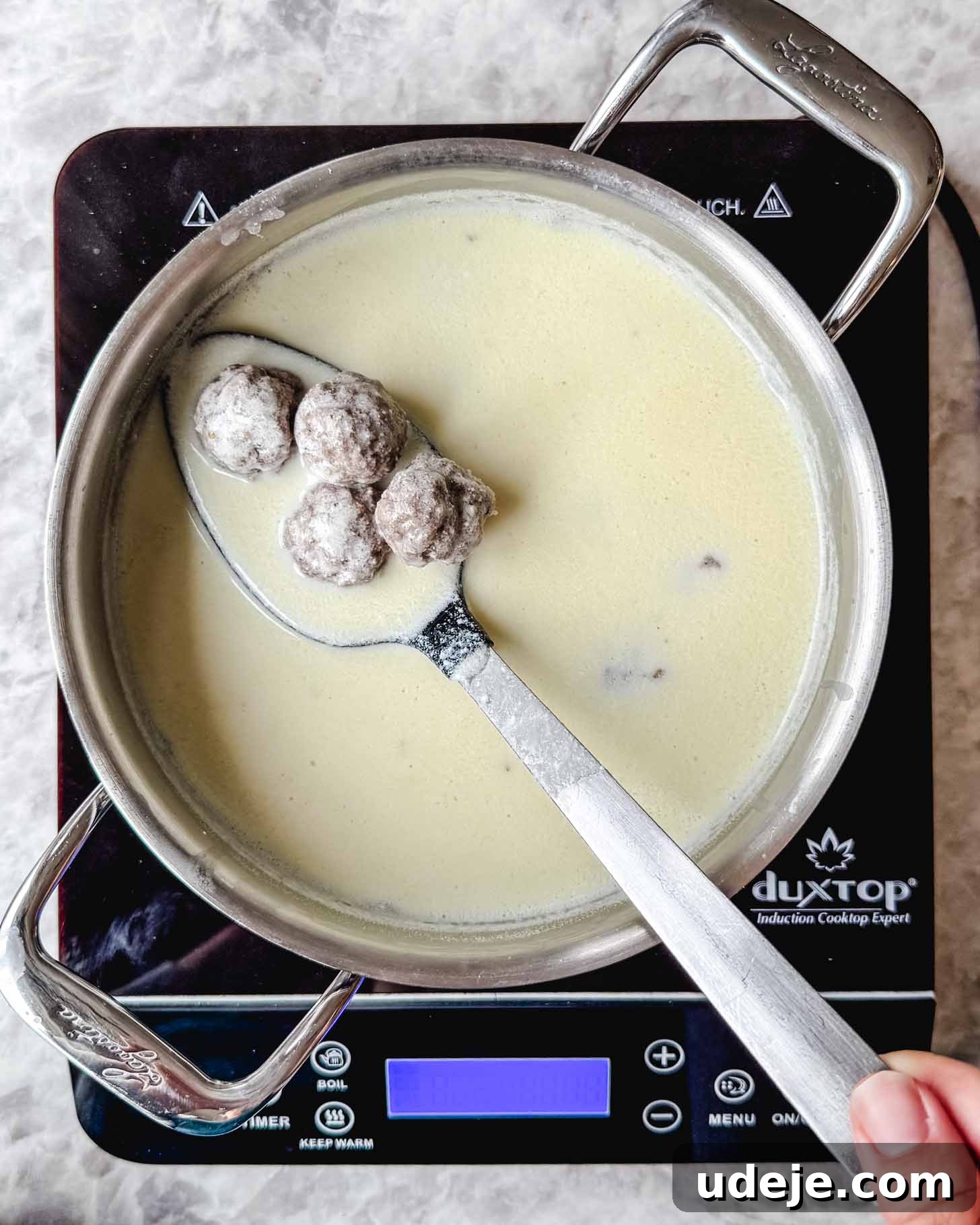 The Ultimate Meatball Soup with Yogurt and Pasta 6 Steel pot with a creamy off white liquid and small brown meatballs in a spoon being held by a hand on a marble background.