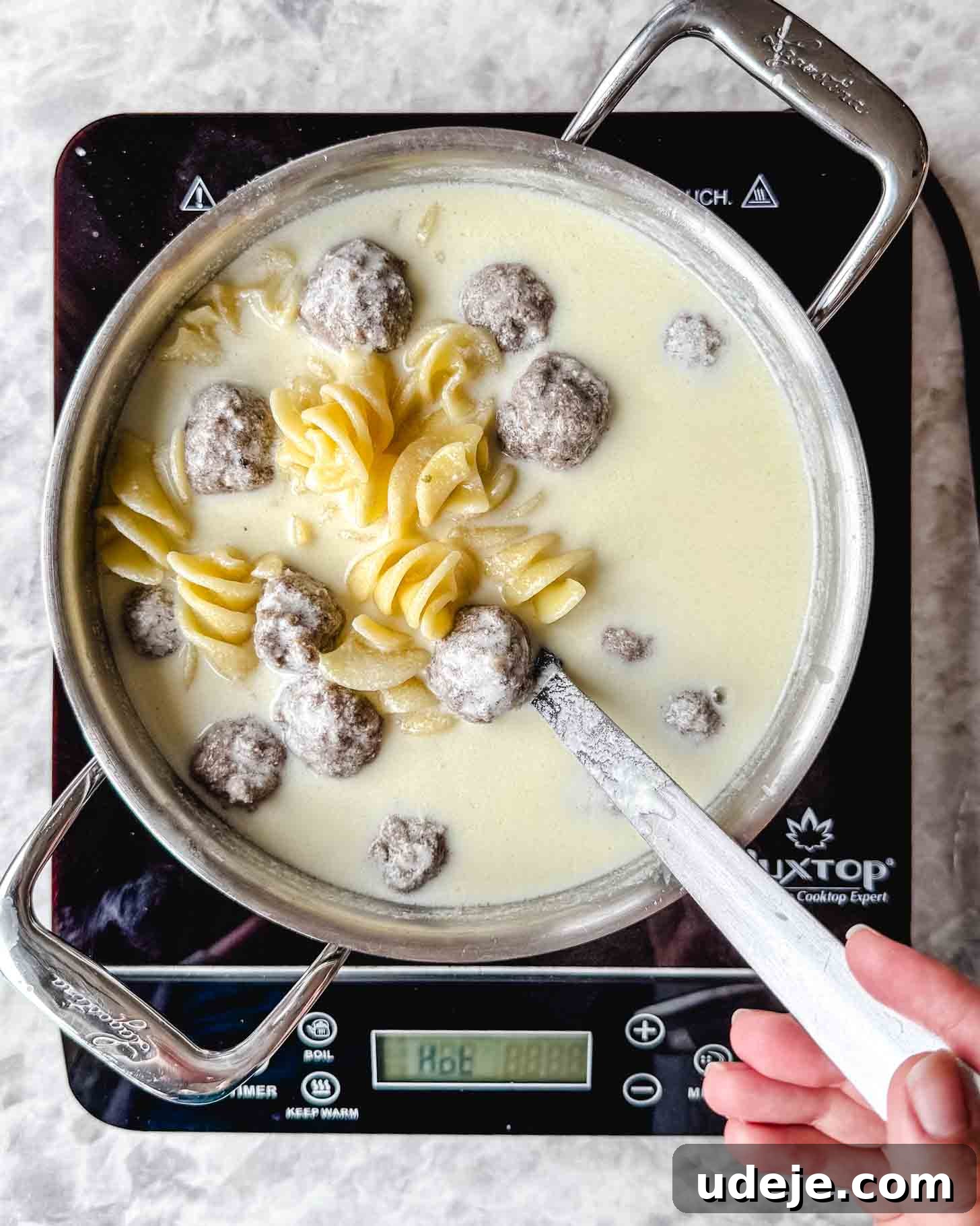 The Ultimate Meatball Soup with Yogurt and Pasta 7 Steel pot with a creamy off white liquid, pasta spirals and small brown meatballs in a spoon being held by a hand on a marble background.