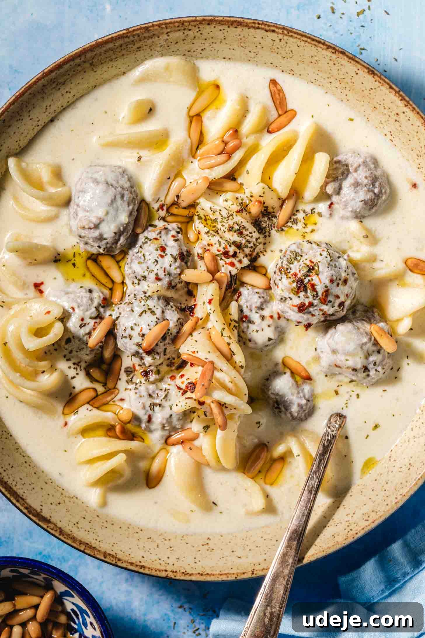 The Ultimate Meatball Soup with Yogurt and Pasta 8 A beige speckled bowl filled with a creamy off-white soup. Small, brown meatballs and spiral-shaped white pasta are visible, all generously topped with golden toasted pine nuts. A metal spoon is placed in the bowl, which sits on a blue background. A blue tea towel is neatly folded in the corner, and a small blue bowl filled with toasted pine nuts is nearby.