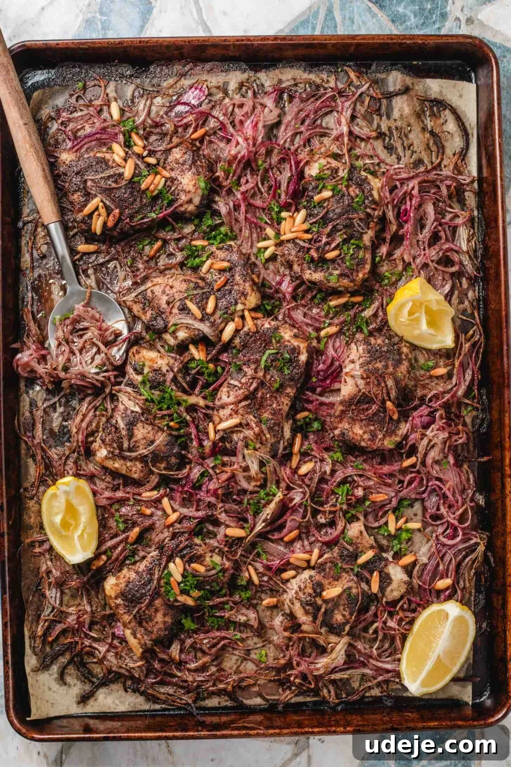 One-Pan Zesty Sumac Chicken and Onions 2 A large sheet pan lined with parchment paper showcasing perfectly cooked chicken thighs, beautifully caramelized red onion slices, and bright lemon wedges. The dish is garnished with toasted pine nuts and fresh parsley, ready to be served.