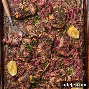 A large sheet pan lined with parchment paper with cooked chicken thighs and cooked slices of red onions, lemon wedges, and a serving spoon. Everything is garnished with toasted pine nuts and parsley.