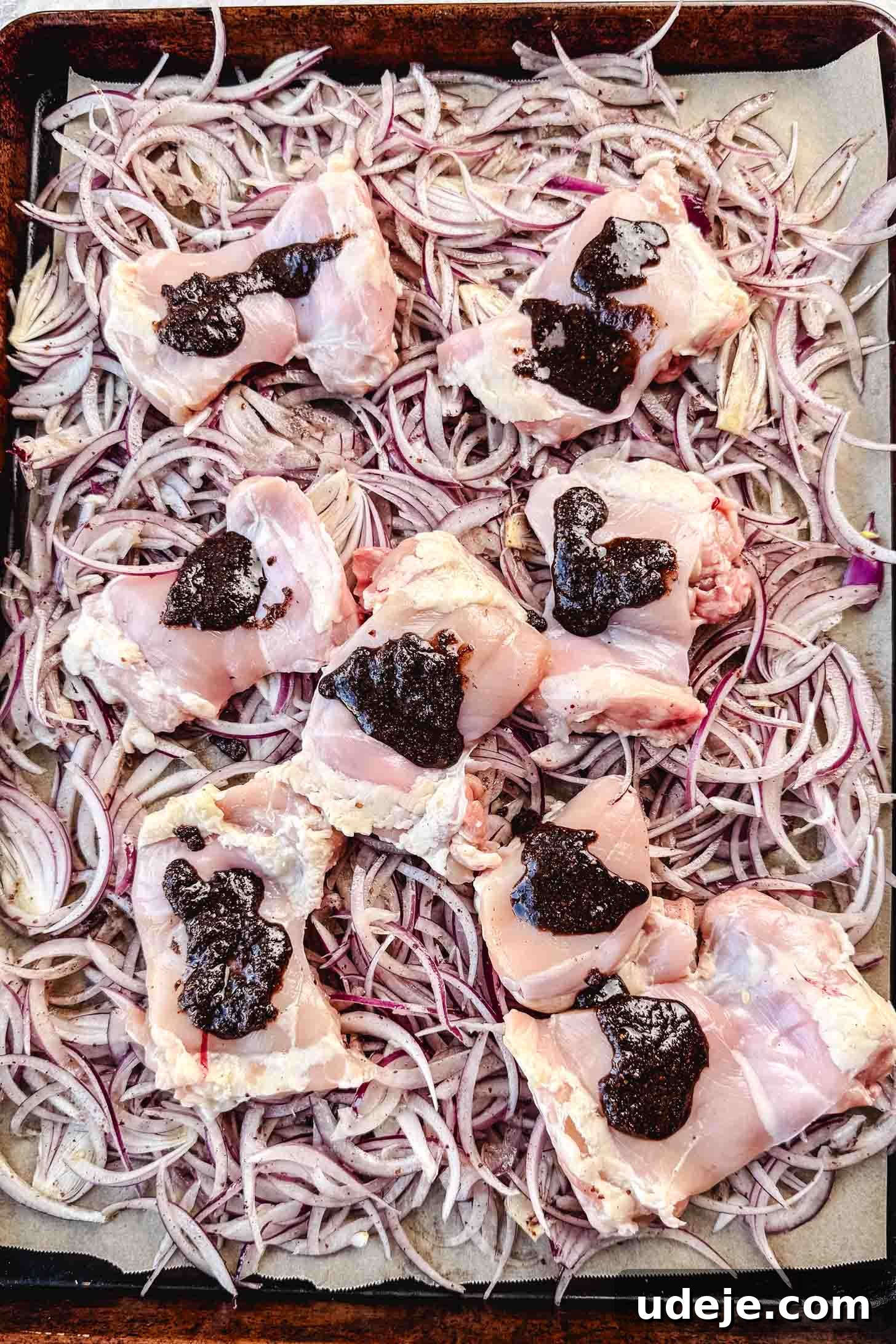 One-Pan Zesty Sumac Chicken and Onions 5 Raw boneless chicken thighs are placed atop the seasoned, thinly sliced red onions on a parchment-lined sheet pan, both awaiting the final spice rub.