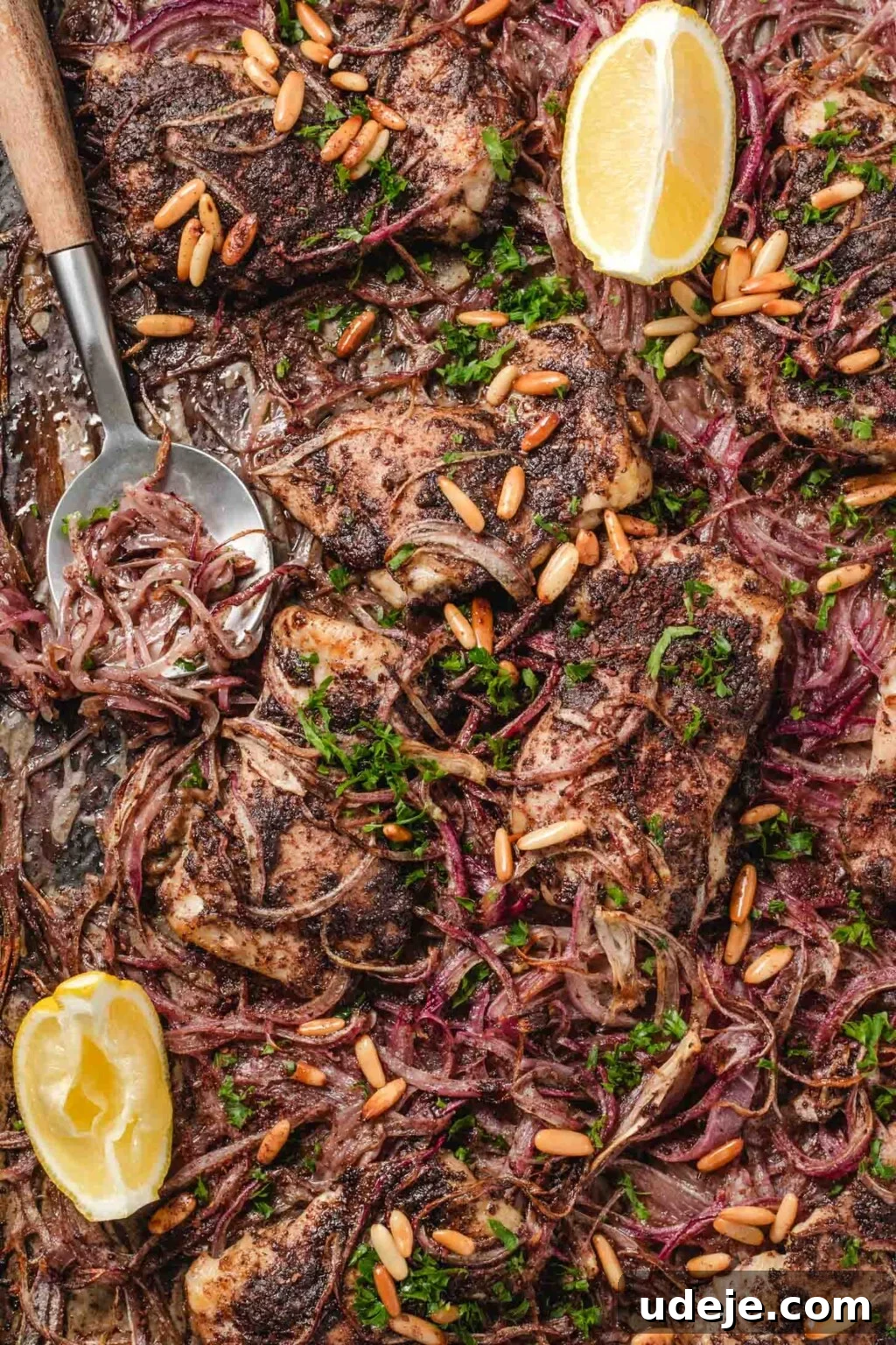 One-Pan Zesty Sumac Chicken and Onions 7 A large sheet pan lined with parchment paper featuring a delicious spread of cooked sumac chicken thighs and perfectly caramelized red onions. The dish is beautifully garnished with fresh lemon wedges, toasted pine nuts, and vibrant parsley, ready for serving.