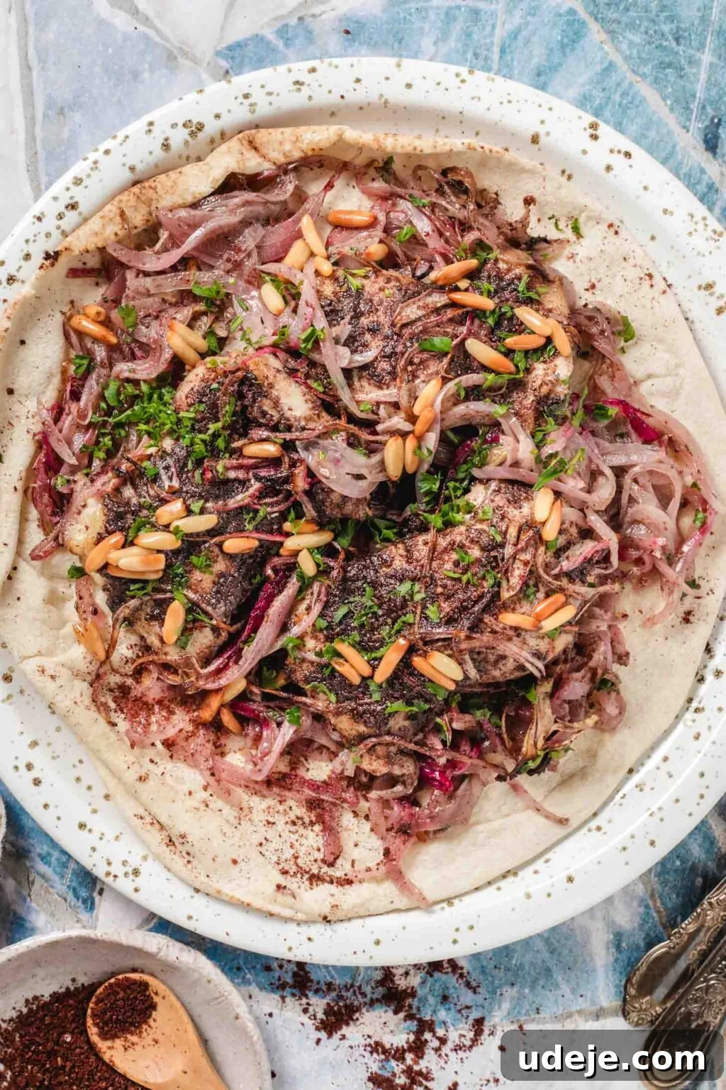 One-Pan Zesty Sumac Chicken and Onions 8 A beautifully plated dinner featuring a large pita flatbread, generously topped with savory baked sumac chicken thighs and an abundance of rich red onions. The dish is elegantly garnished with golden toasted pine nuts and fresh, vibrant parsley.