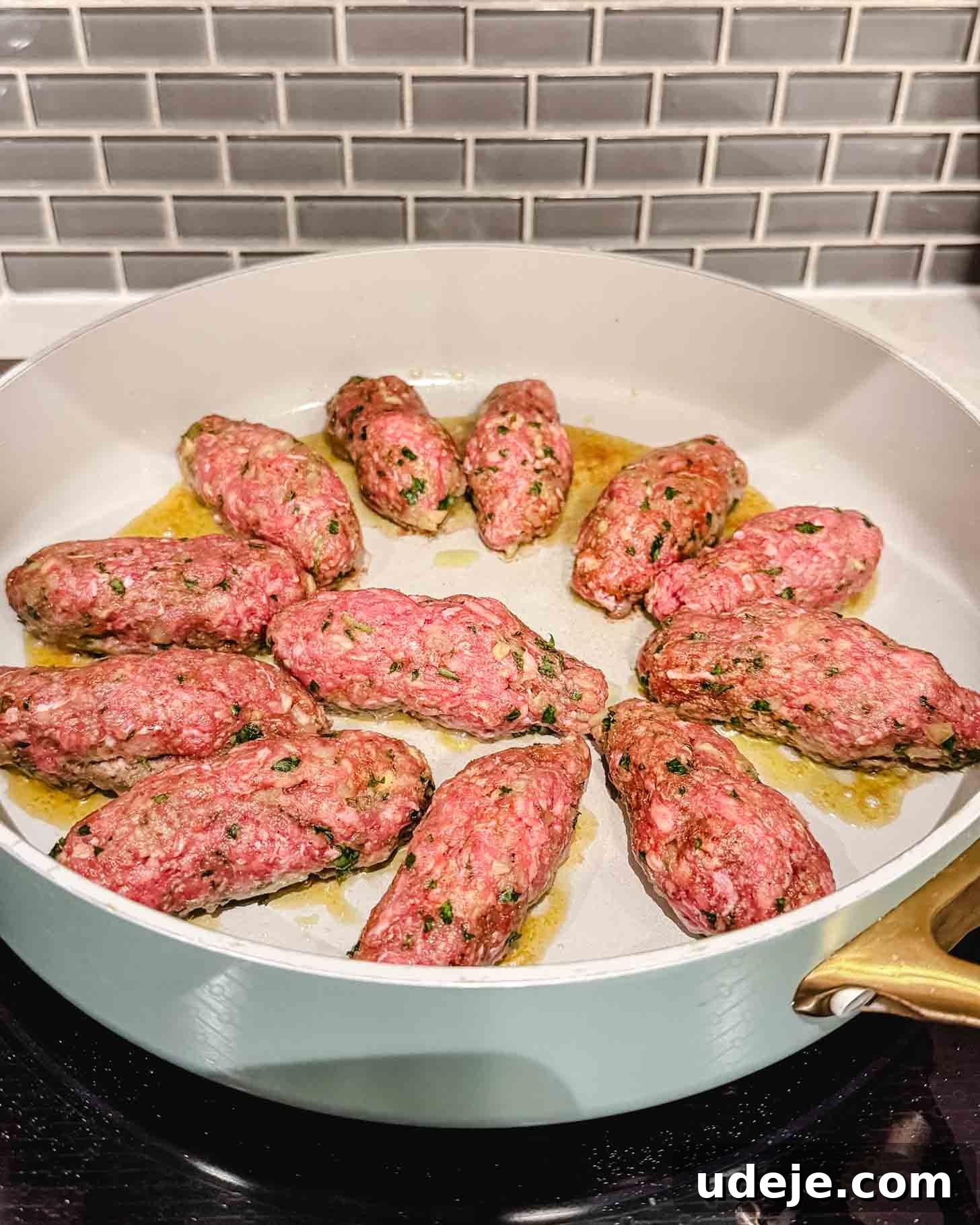 Hearty One-Pot Kofta and Potato Stew 3 Raw, seasoned ground meat shaped into logs is cooking in a shallow white pan with some oil on a stovetop, in front of a gray subway tile backsplash, showing the initial searing process.