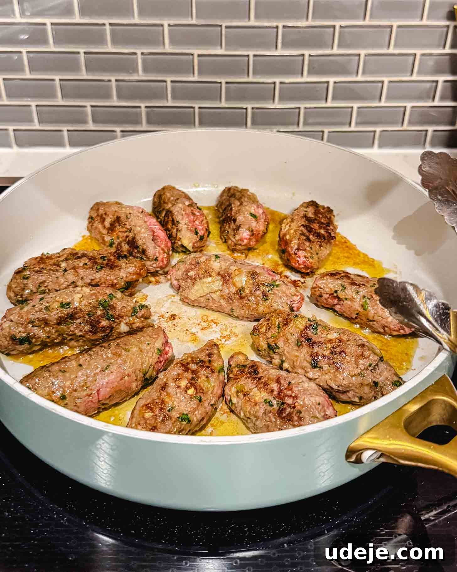Hearty One-Pot Kofta and Potato Stew 4 A white pan with golden handles contains several browned, oval-shaped pieces of ground meat cooking on a stovetop, with a gray tile backsplash in the background. A pair of metal tongs rests on the pan’s edge, indicating the kofta is ready to be removed.