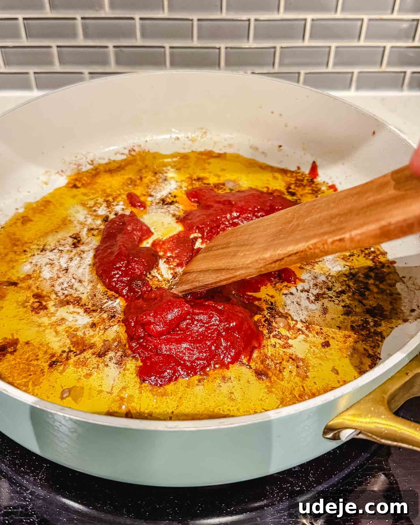 Hearty One-Pot Kofta and Potato Stew 6 A person uses a wooden spatula to stir bright red tomato paste in a white skillet with oil and browned bits on a stovetop, in front of a gray tile backsplash. This step is crucial for building deep flavor in the sauce.