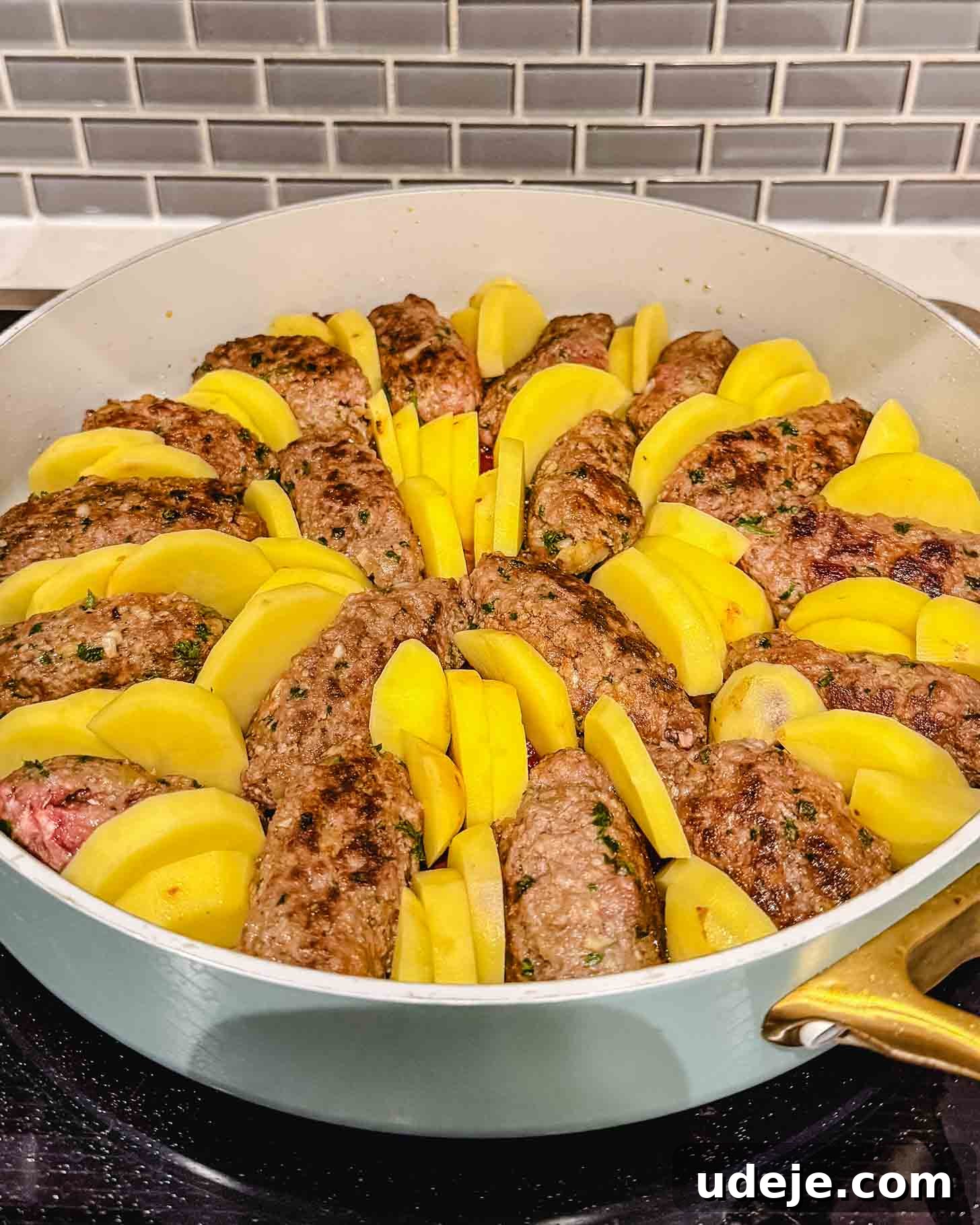 Hearty One-Pot Kofta and Potato Stew 7 A round pan on a stovetop is meticulously arranged with browned oval-shaped meat patties and yellow potato wedges, placed in alternating sections in a circular pattern. The background shows a gray subway tile backsplash, indicating the dish is ready for simmering.