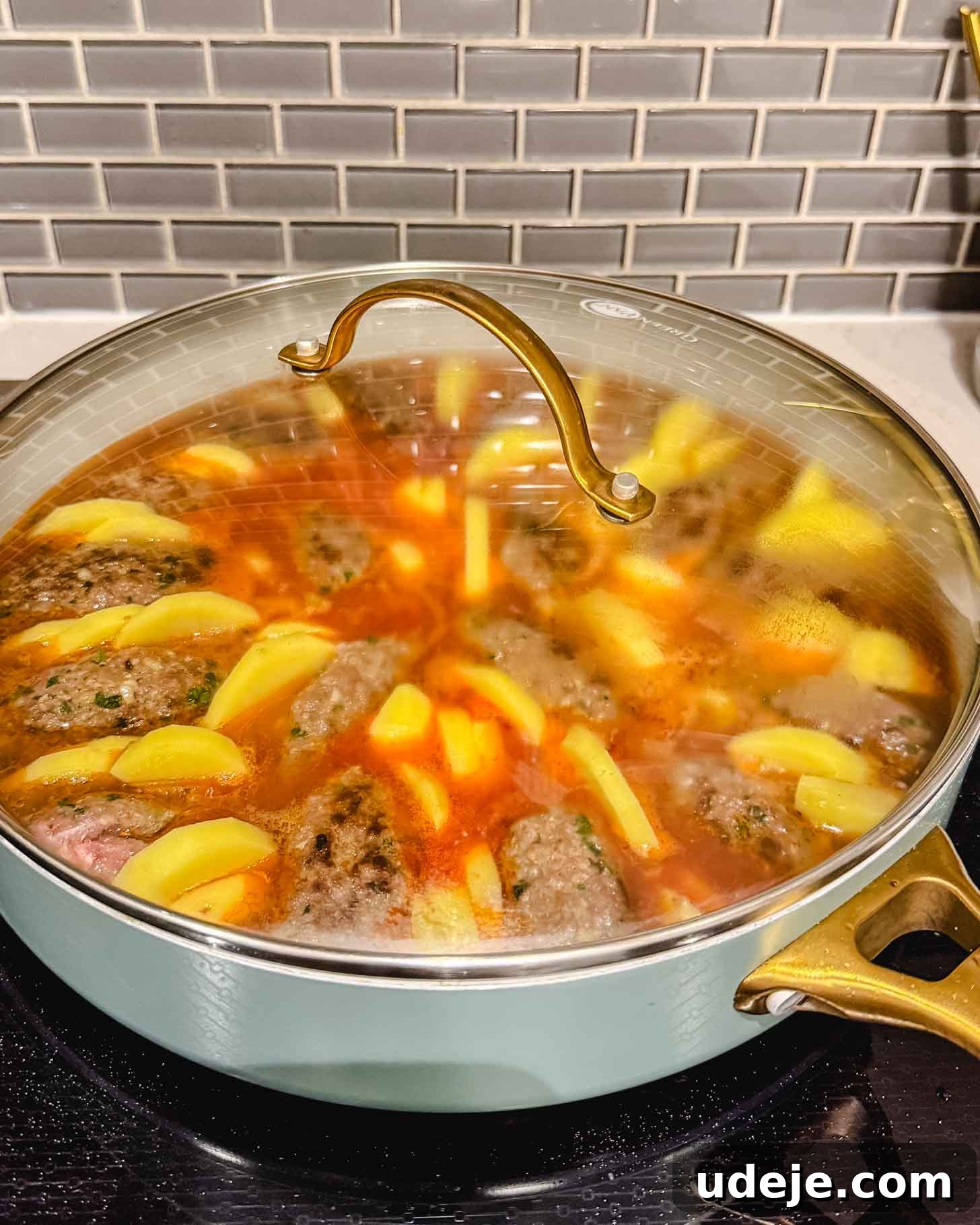 Hearty One-Pot Kofta and Potato Stew 8 A lidded pan on a stovetop contains simmering meat patties, sliced potatoes, and carrots in a reddish broth, with steam gently escaping. The background features a gray tiled backsplash and a white countertop, showing the stew actively cooking.