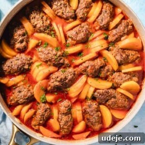 A skillet filled with oval-shaped meat patties and sliced potatoes cooked in a red tomato sauce, garnished with chopped herbs. The dish is on a light blue surface next to a plate of fresh herbs and a glass of water, illustrating the delicious Kofta and Potato Stew.
