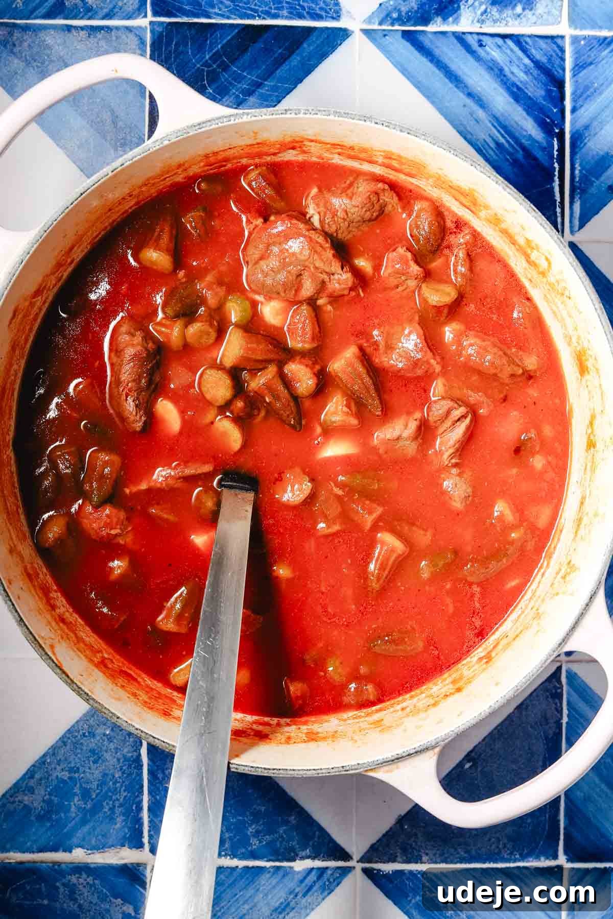Flavorful Middle Eastern Bamya Okra Stew 11 A large white pot showcasing a fully mixed and simmering okra stew, rich with veal cubes, onions, garlic, and tomato puree, stirred with a metal spoon on a blue and white patterned tile background.