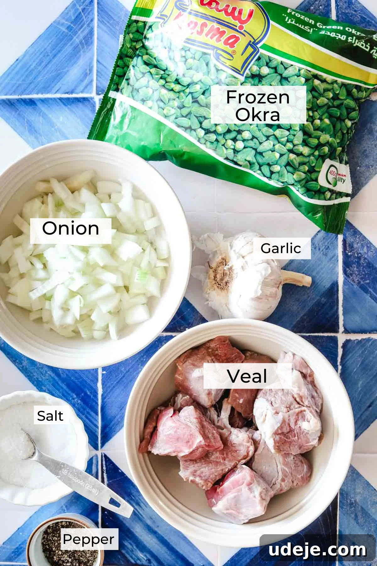 Flavorful Middle Eastern Bamya Okra Stew 5 An assortment of fresh ingredients for okra stew laid out on a blue and white patterned tile. This includes a bag of frozen okra, a white bowl of chopped white onion, a whole head of garlic, a bowl of veal cubes, and small white bowls of salt and black pepper with a metal spoon.