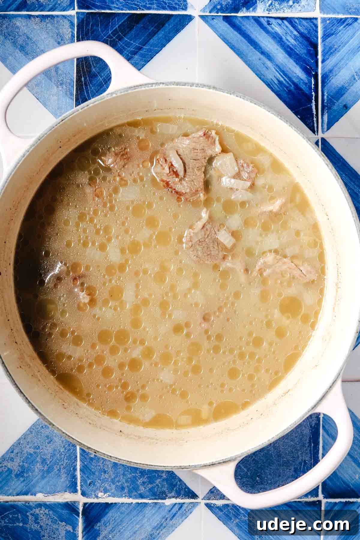 Flavorful Middle Eastern Bamya Okra Stew 6 A large white pot with handles containing a rich broth, translucent onions, and succulent veal pieces, all simmering on a blue and white patterned tile background.