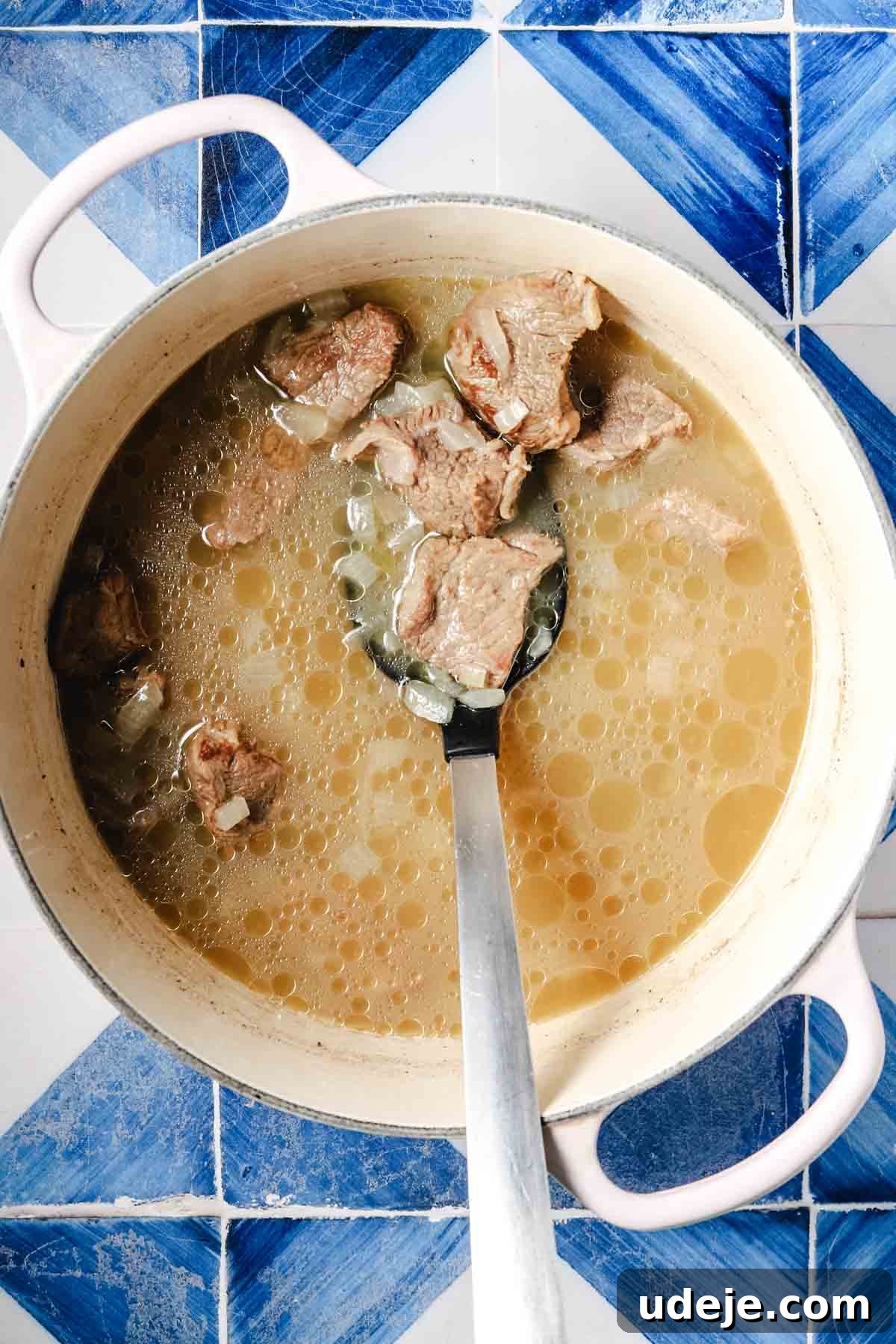 Flavorful Middle Eastern Bamya Okra Stew 7 A large white pot with handles showcasing a simmering mixture of broth, onions, and veal pieces, with a metal spoon resting within, set against a blue and white patterned tile background.
