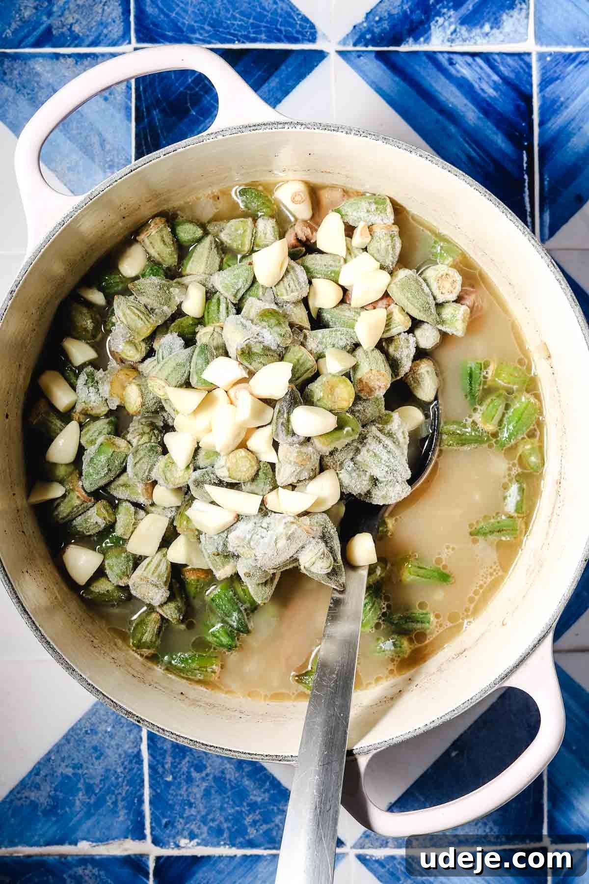 Flavorful Middle Eastern Bamya Okra Stew 9 A large white pot filled with a vibrant mixture of broth, tender veal cubes, finely chopped onions, roughly chopped garlic, and frozen okra, all ready for the next stage of cooking with a metal spoon.
