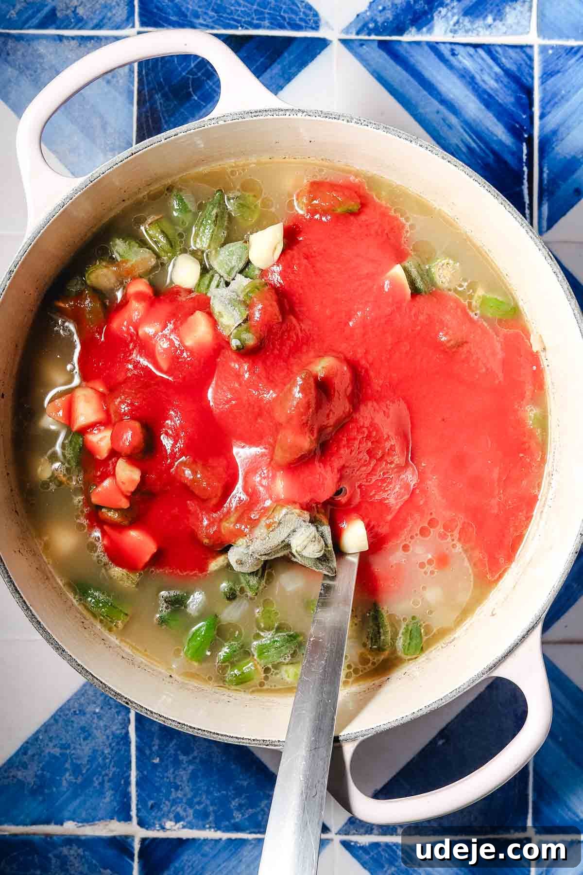 Flavorful Middle Eastern Bamya Okra Stew 10 A large white pot containing broth, veal cubes, chopped onions, garlic, and frozen okra, now with added tomato puree, gently stirred with a metal spoon, showcasing the rich base of the stew.