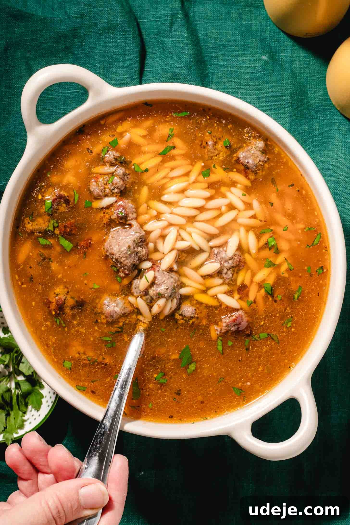 Kofta-Style Meatball Orzo Broth 3 A hand holding a spoon dips into a white bowl of Brothy Meatball and Orzo Soup, rich with tender meatballs, delicate orzo pasta, and vibrant chopped herbs, all set against a lush green tablecloth.