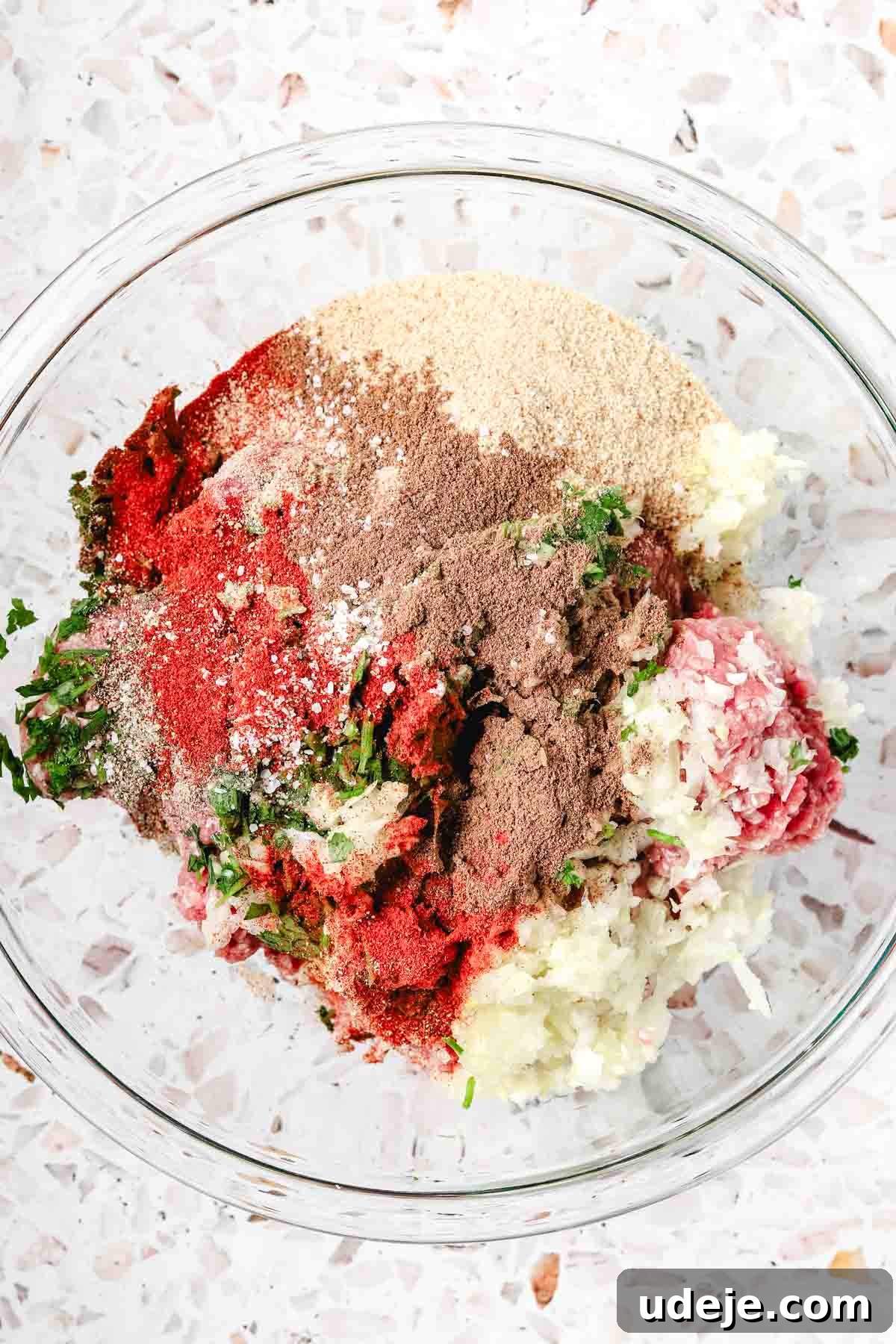 Kofta-Style Meatball Orzo Broth 4 A medium-sized glass bowl holds a vibrant mixture of ground beef, finely diced onions, fresh parsley, and a selection of aromatic spices, all resting on a stylish terrazzo patterned background.