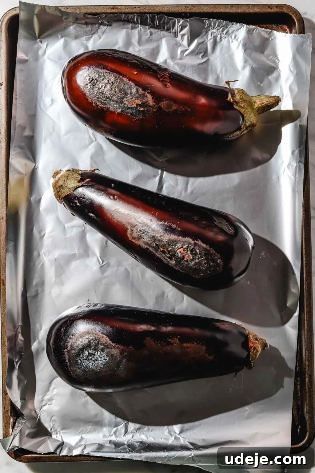 Charred Eggplant Baba Ghanoush 4 Three large eggplants resting on a foil-lined baking sheet, lightly roasted on their top side, showcasing the beginning of the roasting process.