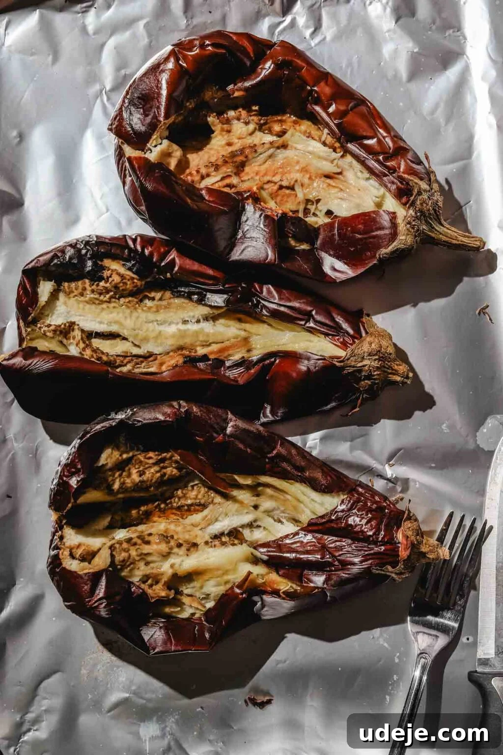Charred Eggplant Baba Ghanoush 6 Three large roasted eggplants, split open lengthwise on a foil-lined baking sheet, with a fork and knife visible in the corner, ready for the flesh to be scooped out.