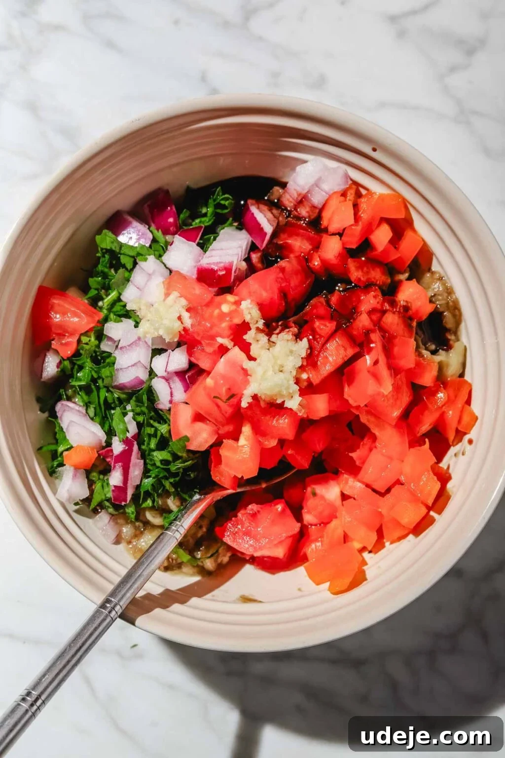 Charred Eggplant Baba Ghanoush 10 Mashed eggplant in a small beige bowl, brimming with a colorful mix of chopped parsley, garlic, red onions, red pepper, and tomatoes, with a spoon nestled within, all on a light marble background.