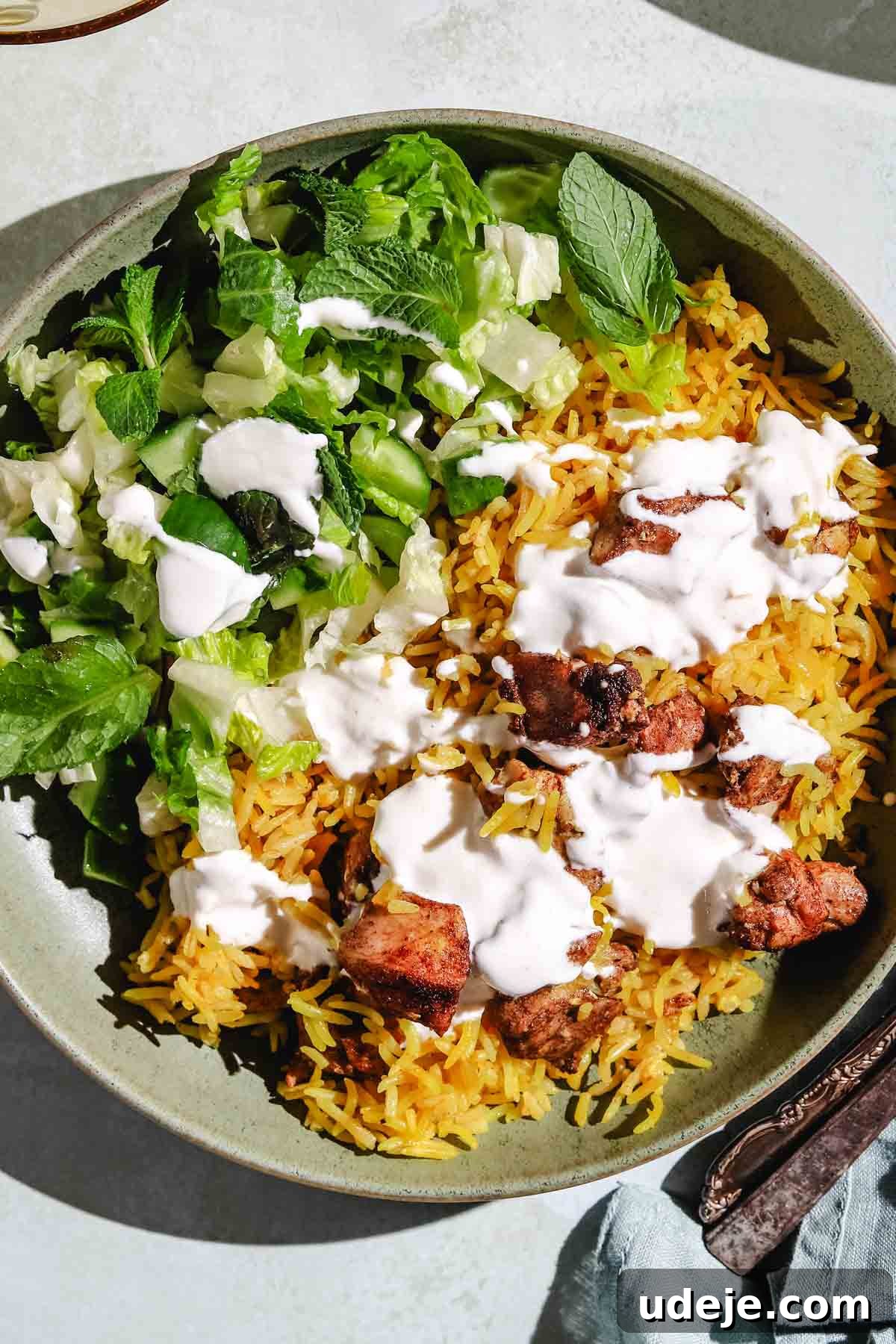 Shawarma rice consisting of yellow rice with chicken pieces in a plate, drizzled with white sauce and with a green salad on the side.