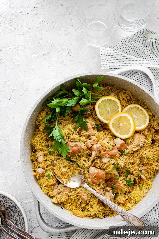 One-Pot Shawarma Rice with Creamy Garlic Sauce 3 A warm skillet filled with golden yellow shawarma rice and tender chicken pieces, beautifully garnished with fresh parsley and two lemon slices. A serving spoon is ready, with elegant glassware and napkins arranged on a light-textured surface, highlighting a comforting meal.