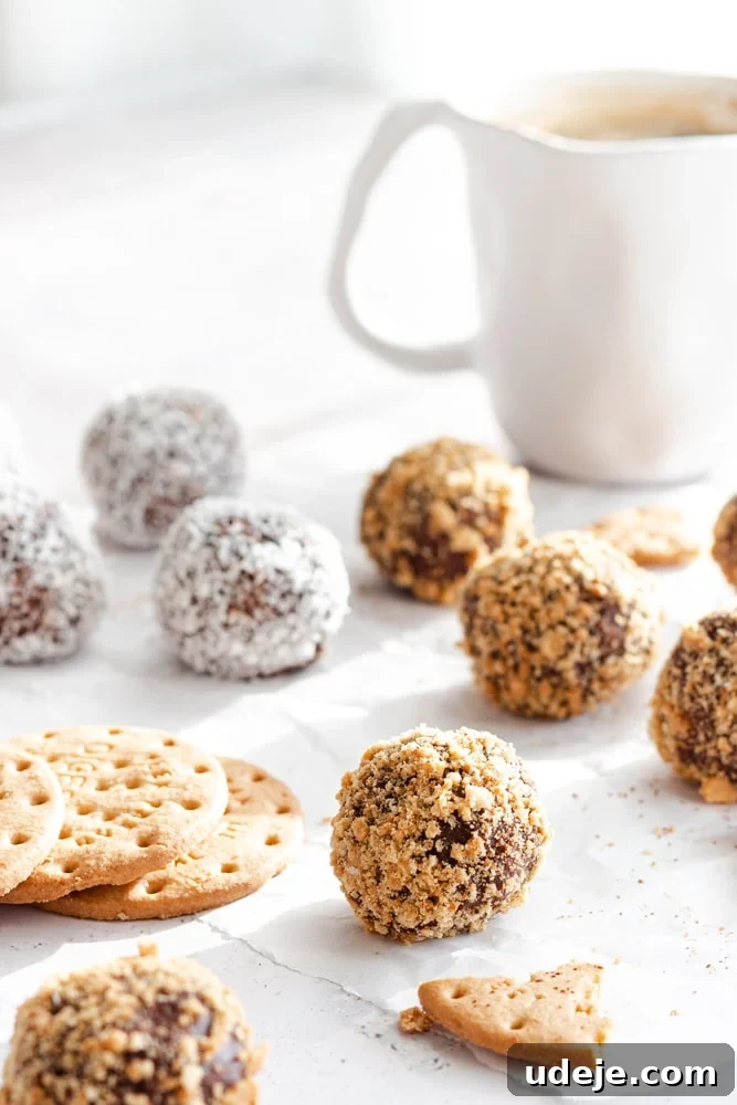 No-Bake Three-Ingredient Chocolate Cookie Bites 5 Close-up of chocolate biscuit balls garnished with crushed biscuits and shredded coconut, artfully arranged on parchment paper next to a white coffee mug. Whole digestive biscuits are visible in the background.