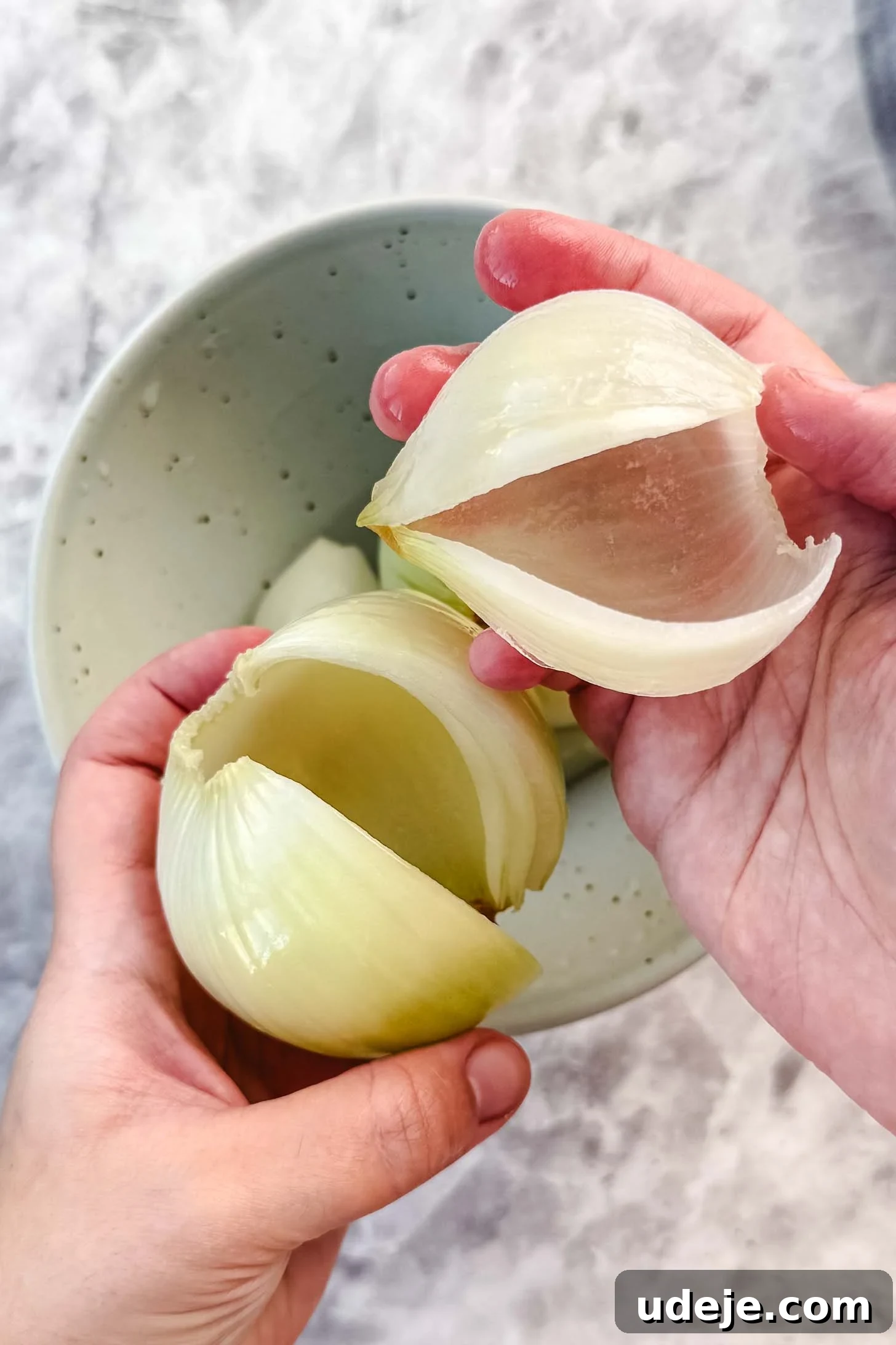 Iraqi Dolma: Stuffed Vegetable Delights 11 Peeled and slit onion being held by two hands, with one hand holding a layer of the onion over a blue speckled bowl on a marble background.