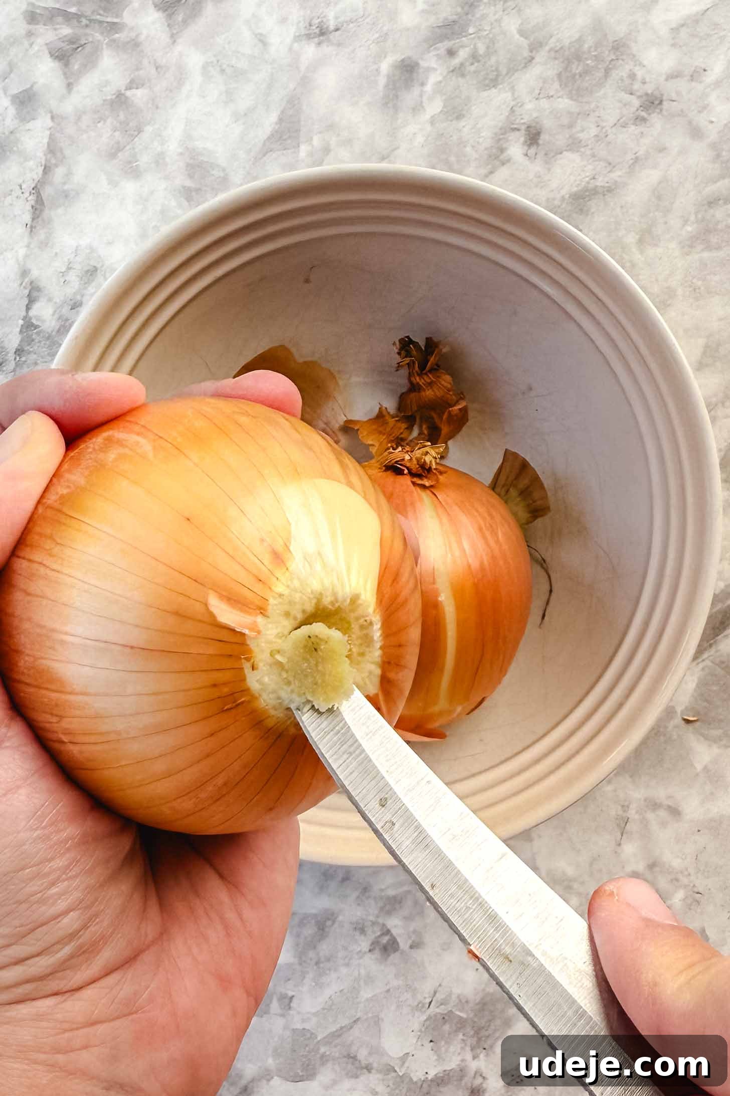 Iraqi Dolma: Stuffed Vegetable Delights 9 Onion being held and the bottom being cut off with knife over a white bowl on a marble background.