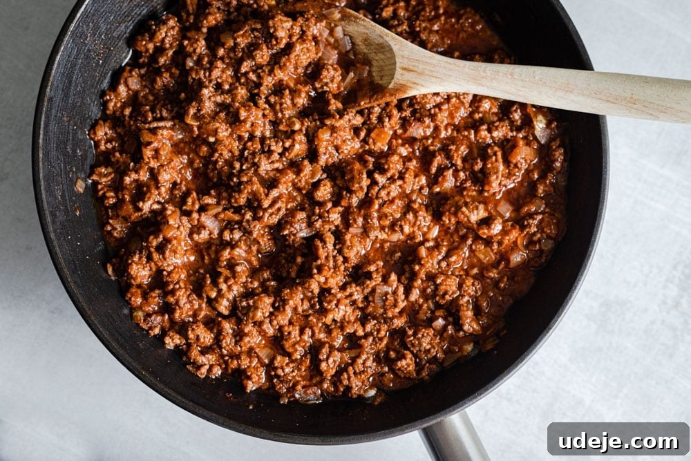 Creamy Bechamel Macaroni Bake 4 Sizzling ground meat sauce cooking in a black pan, being stirred with a wooden spoon, releasing its fragrant spices.
