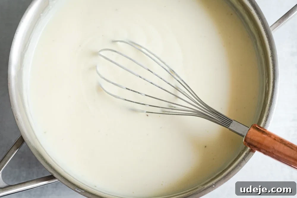 Creamy Bechamel Macaroni Bake 5 A shiny silver pan containing creamy, white Bechamel sauce, with a whisk resting inside, showing its smooth texture.