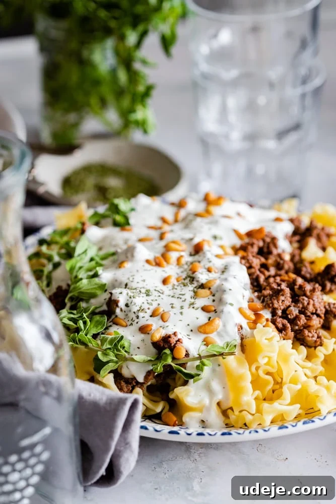 Quick Arabic Pasta with Zesty Yogurt Sauce 6 A beautifully plated dish of Arabic pasta, featuring layers of pasta, spiced ground beef, and a creamy yogurt sauce, lavishly topped with golden toasted pine nuts and vibrant fresh herbs. A clear glass and jug are blurred in the background, suggesting a refreshing drink.